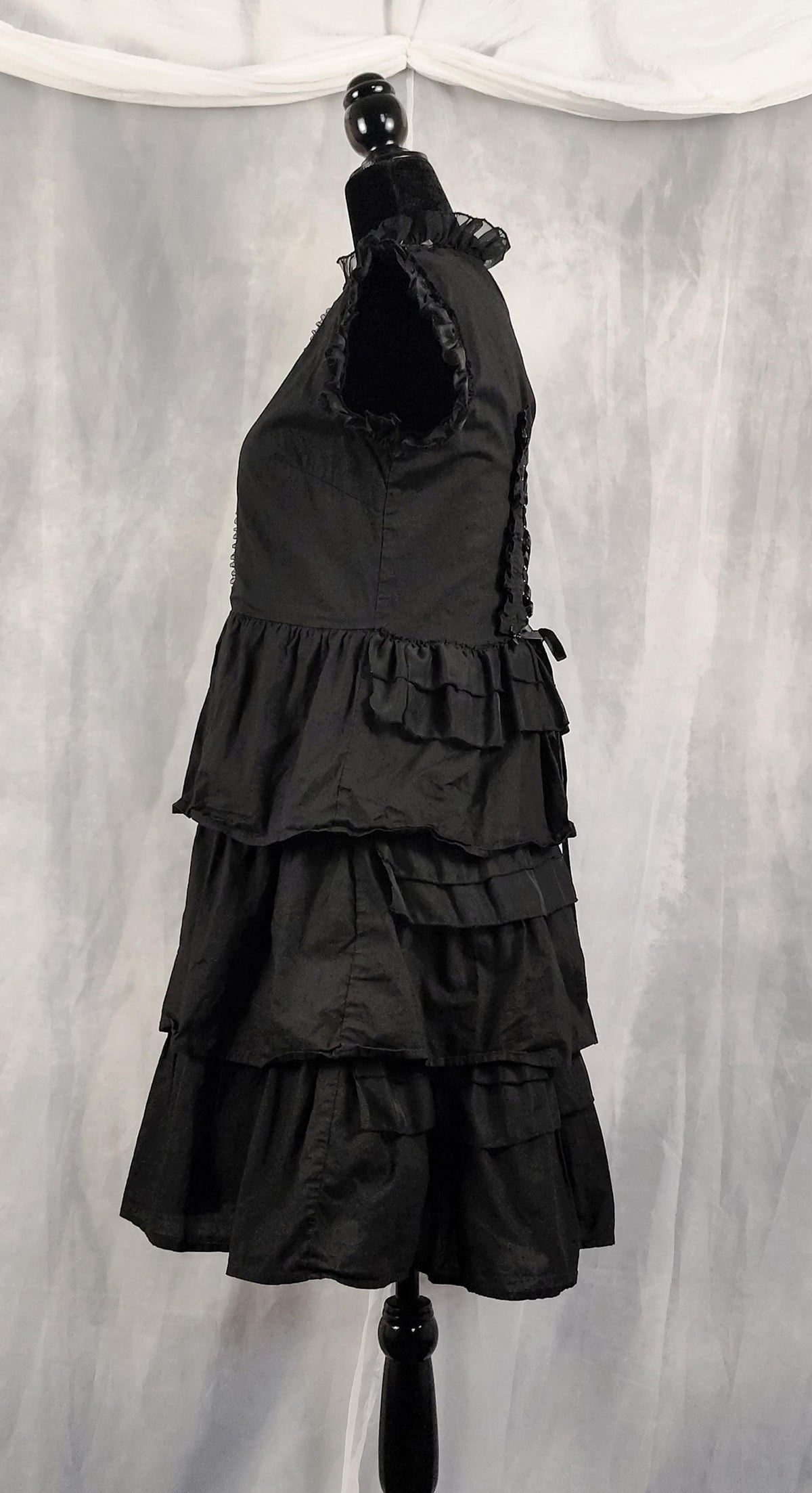 Side view of a black sleeveless tiered dress displayed on a black mannequin against a gray draped fabric backdrop.