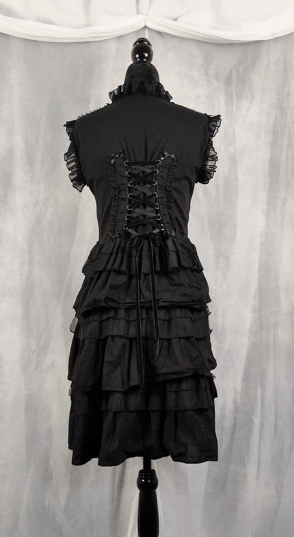 Back view of a black Victorian-style ruffled dress displayed on a black dress form mannequin, showing the back view with lace-up detailing and tiered skirt, set against a gray draped fabric backdrop.