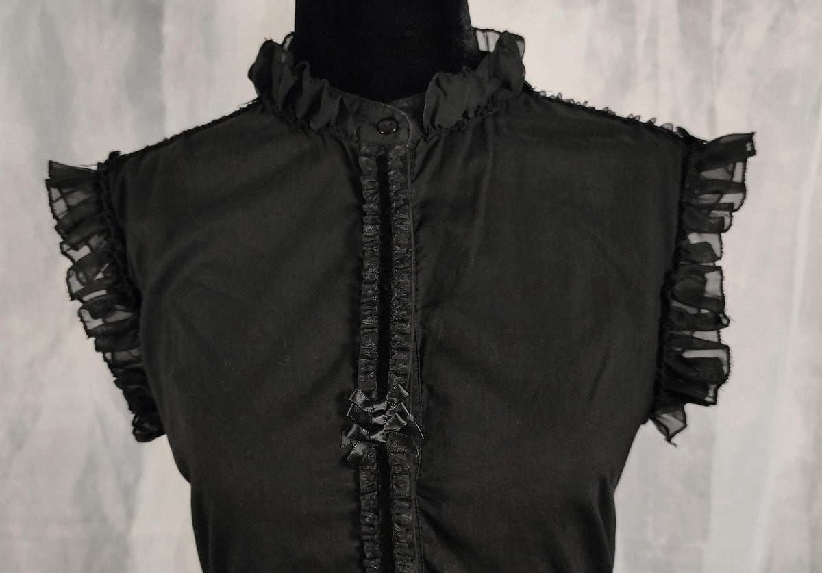Close-up of a black sleeveless dress with sheer ruffled cap sleeves and a ruffled button placket, displayed on a black mannequin torso against a light gray textured backdrop.