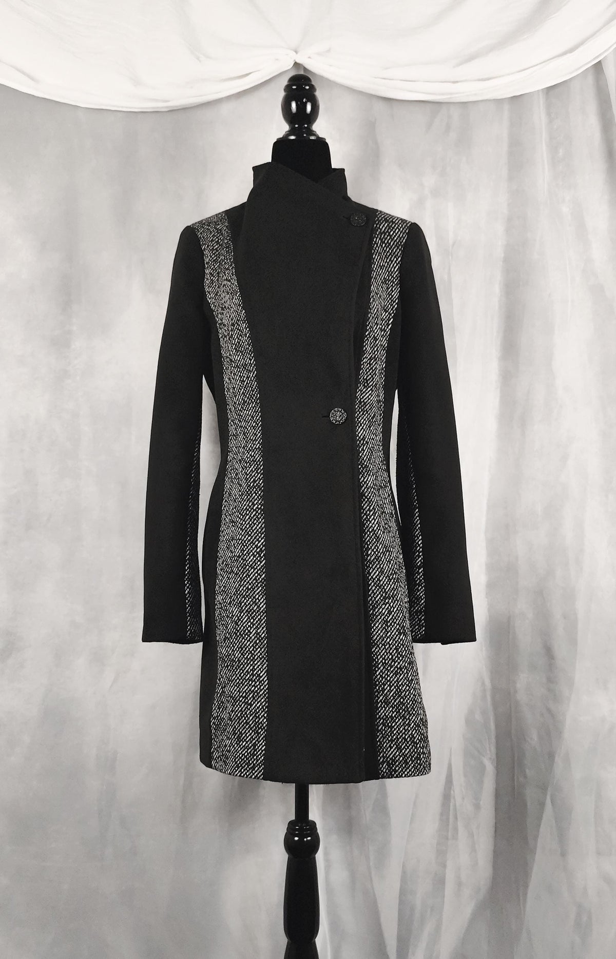 Front view of a black longline coat with textured gray and white patterned side panels, displayed on a black dress form mannequin against a draped gray fabric backdrop.