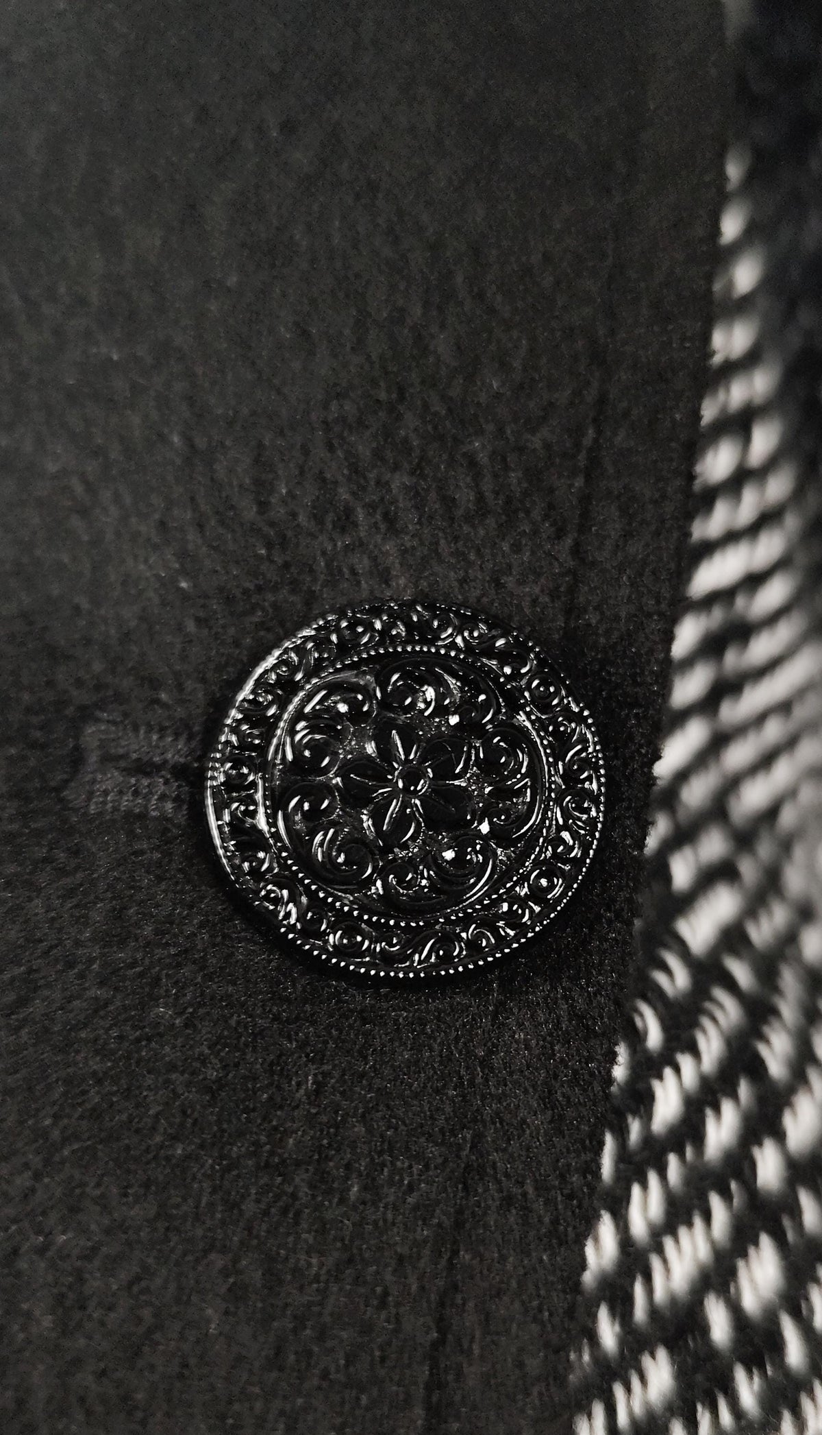 A close-up of a black ornate round button with floral detailing, set on a black textured fabric next to a black and white patterned ribbon.