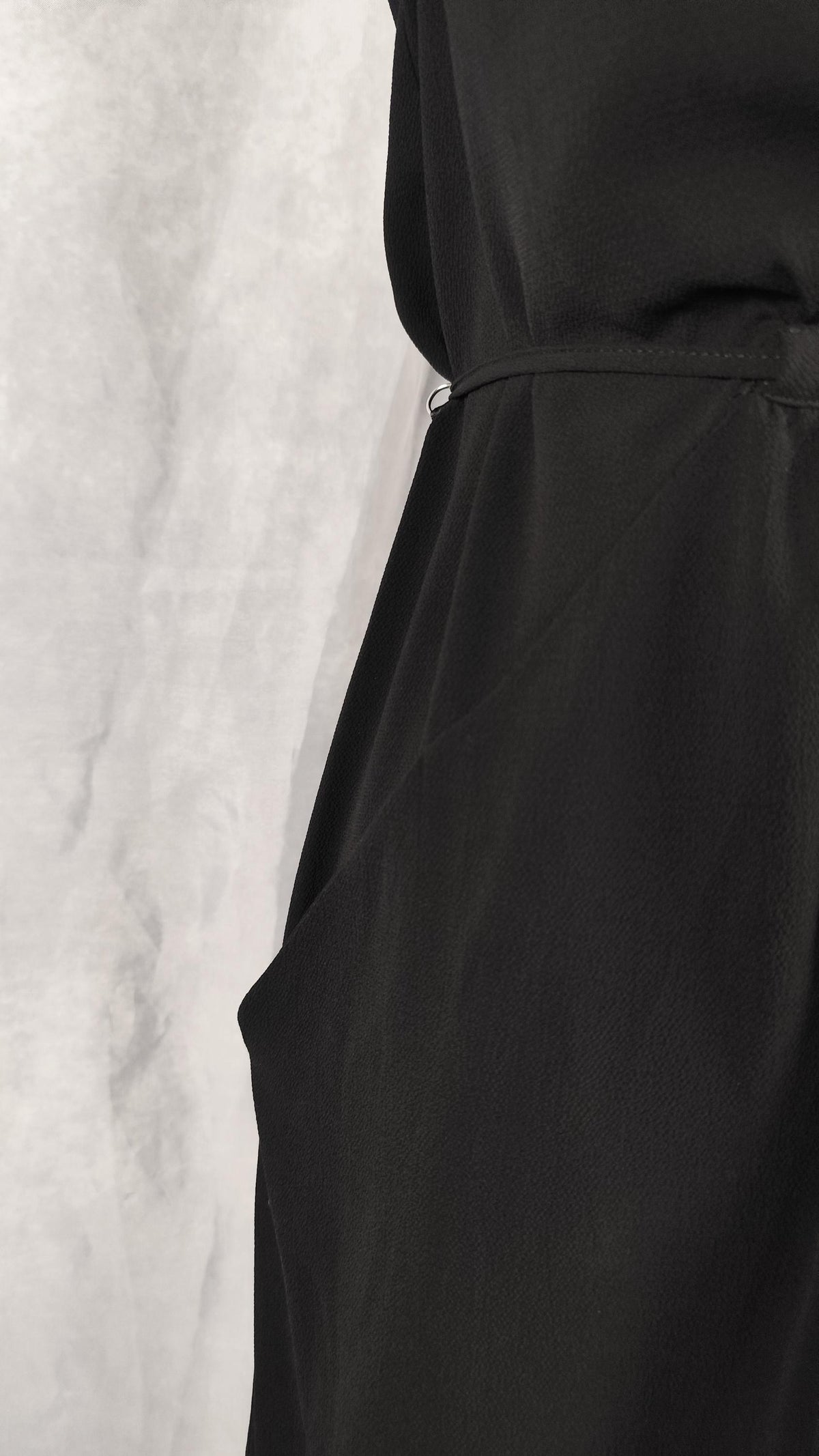 Close-up of a black dress showing the waistband and side pocket against a light gray textured backdrop.