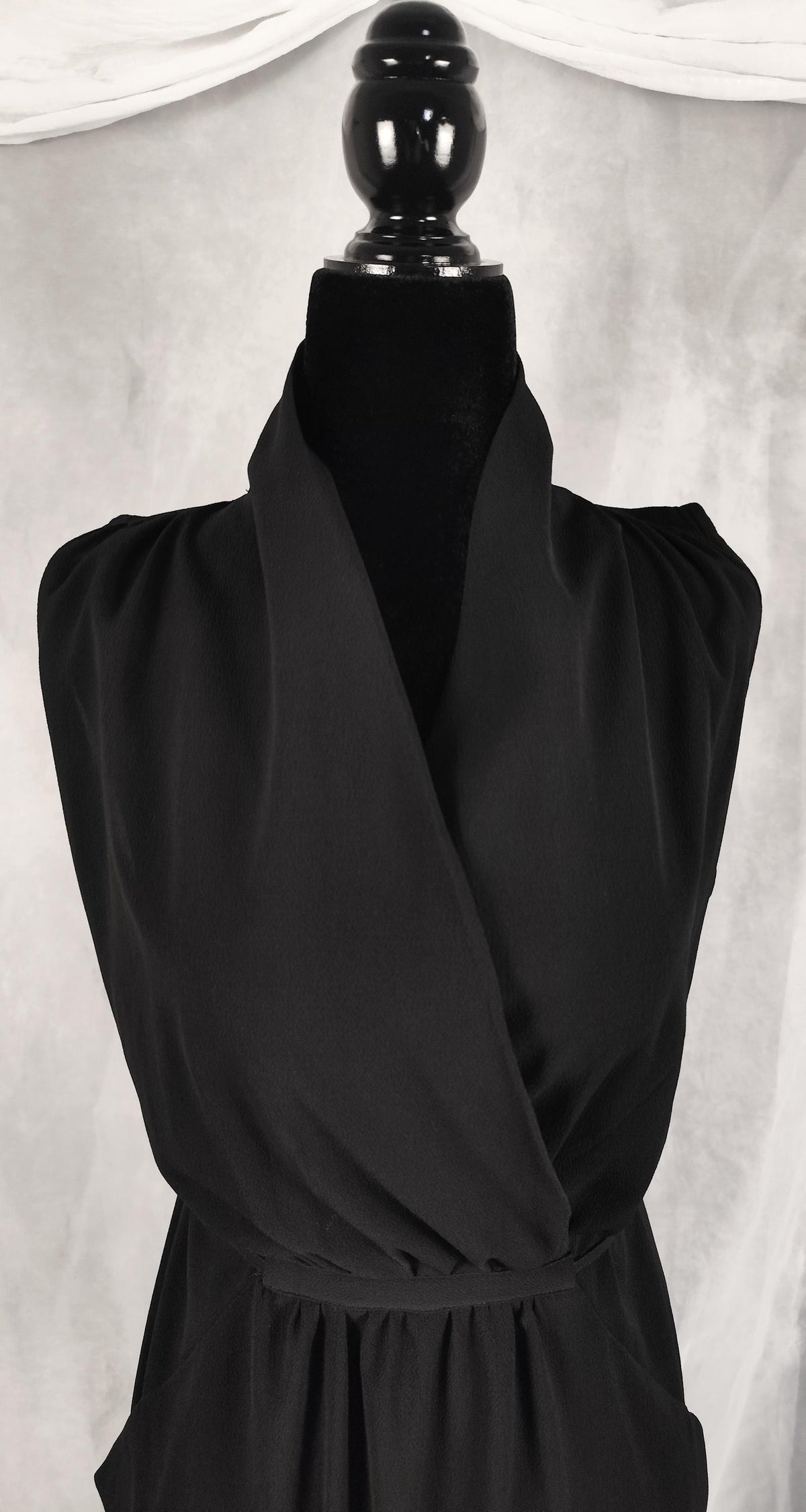 A black sleeveless wrap-style dress displayed on a black mannequin torso against a light gray draped fabric backdrop.
