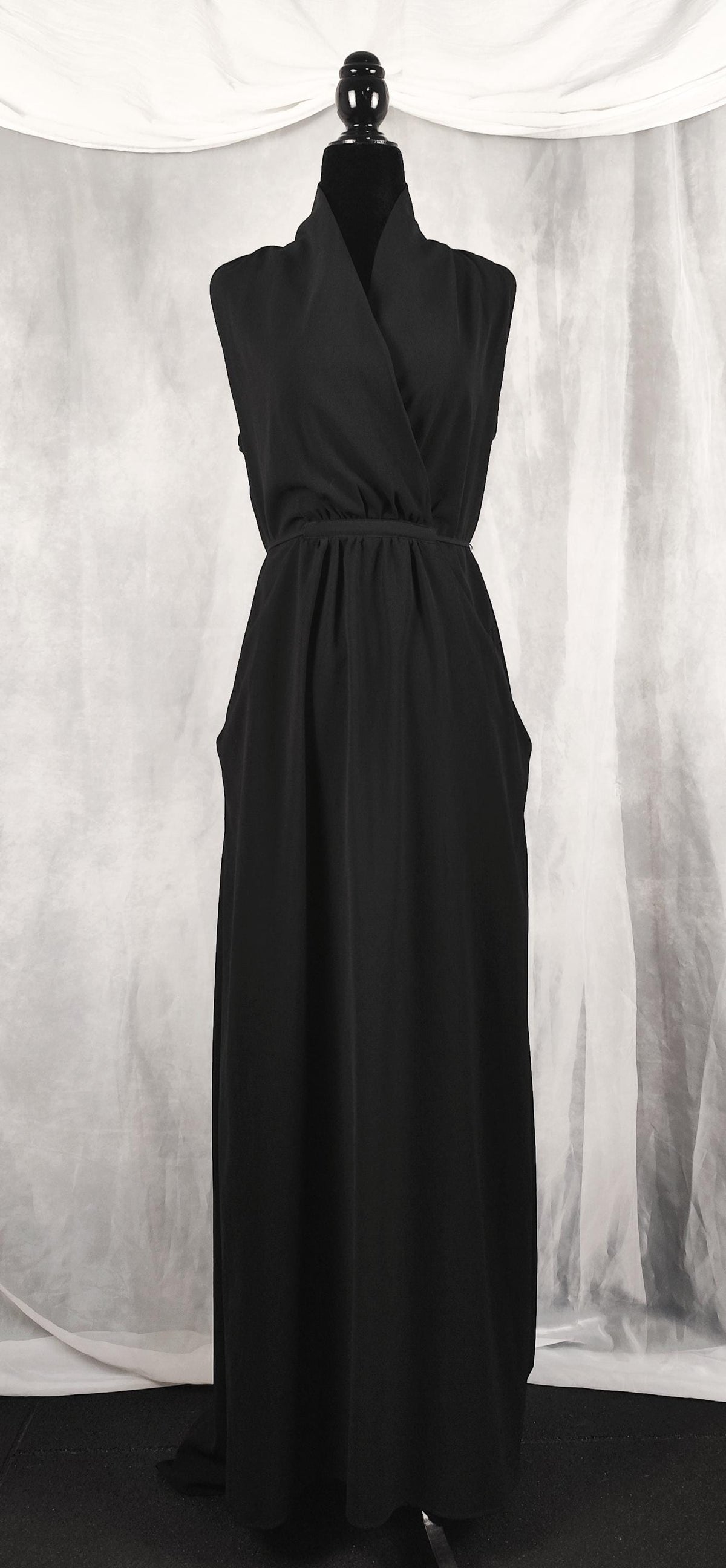 Front view of a sleeveless black wrap-style maxi dress displayed on a black mannequin against a draped gray fabric backdrop.