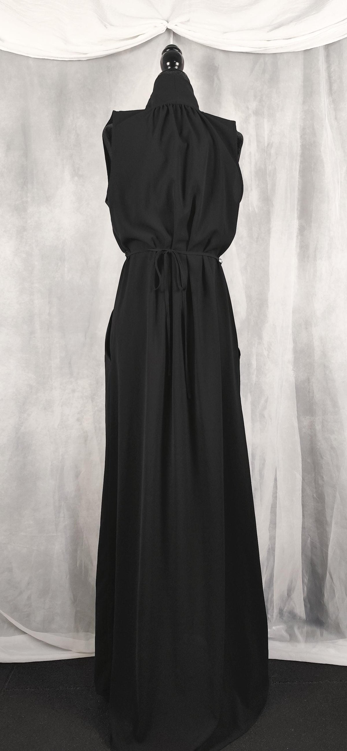 Back view of a sleeveless black maxi dress with a high gathered neckline and a drawstring waist, displayed on a black mannequin against a gray draped fabric backdrop.