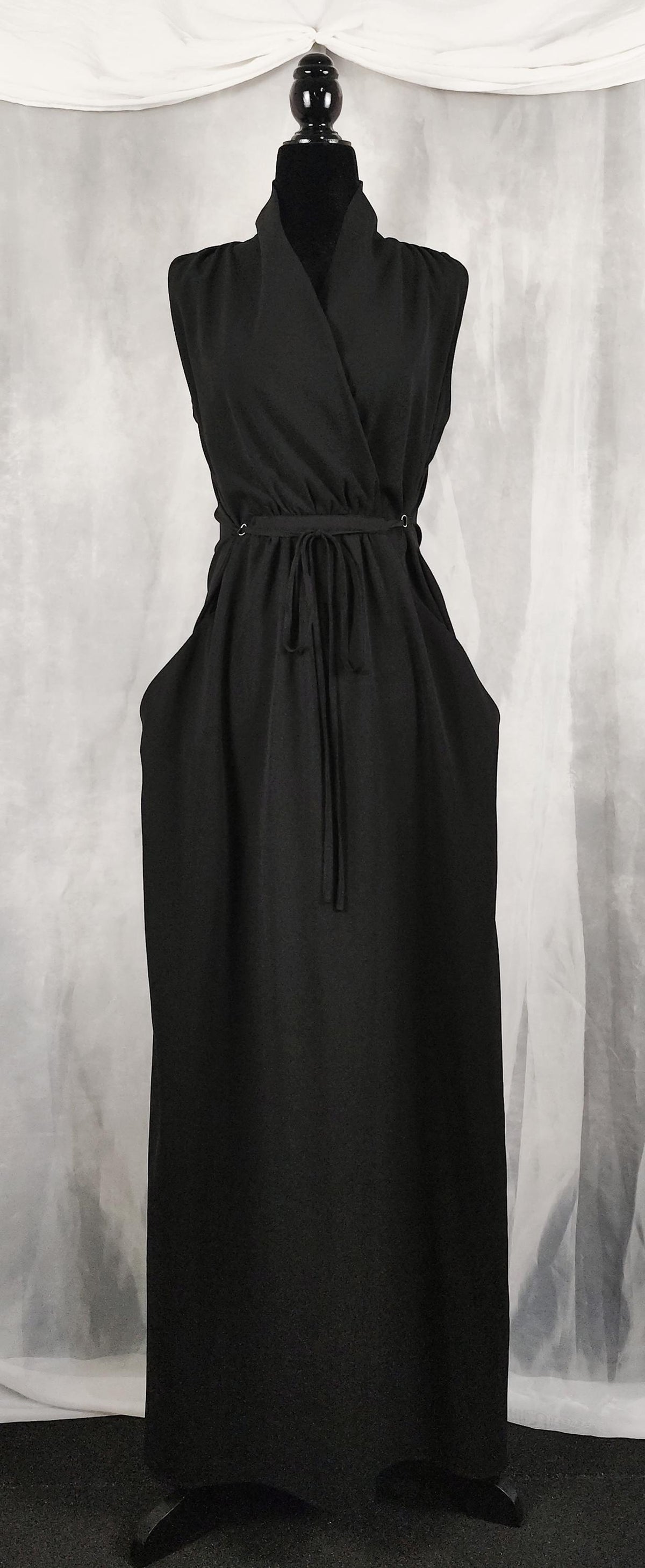 Front view of a sleeveless black maxi dress displayed on a mannequin. The dress features a crossover V-neckline, an adjustable drawstring waist tie, and a long, flowing skirt with side pockets