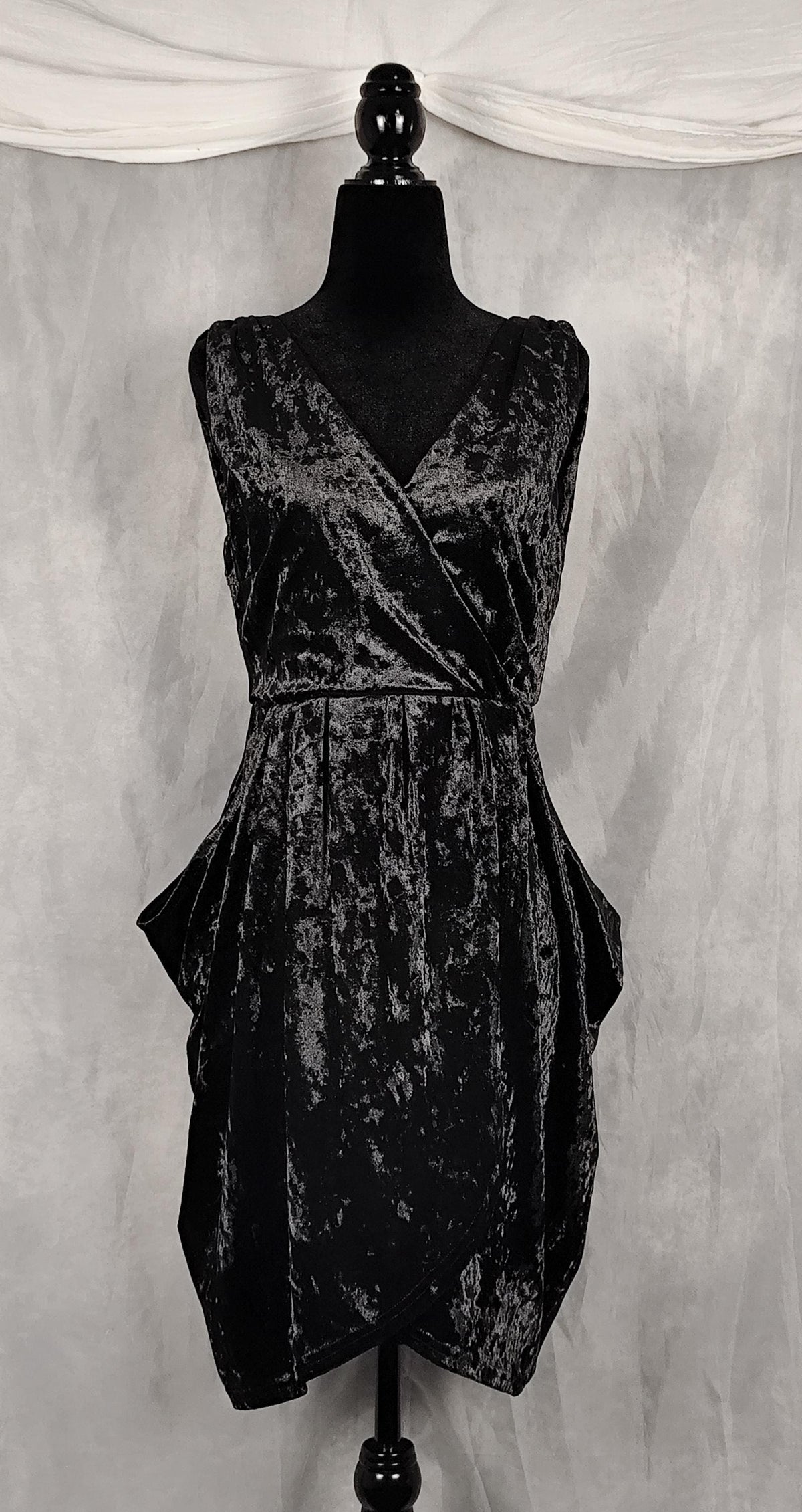 A sleeveless black crushed velvet wrap-style dress displayed on a black mannequin against a gray draped fabric backdrop.