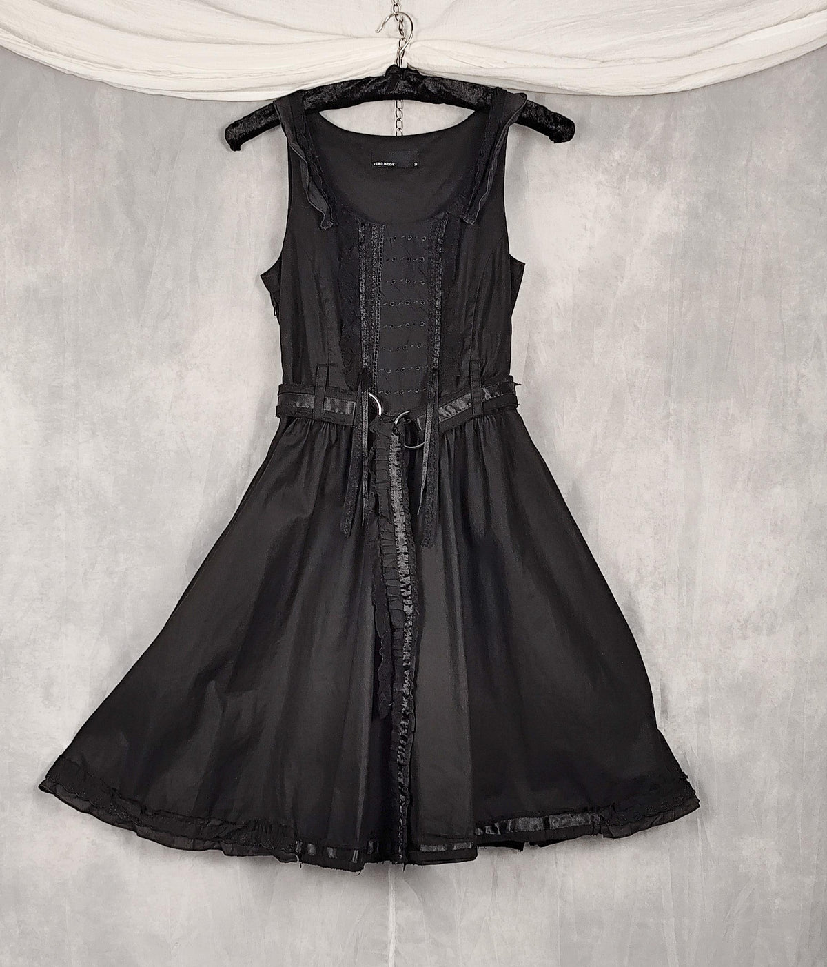 Front view of a black sleeveless dress with a structured corset-style bodice, and matching belt, hanging on a black hanger against a textured gray backdrop.