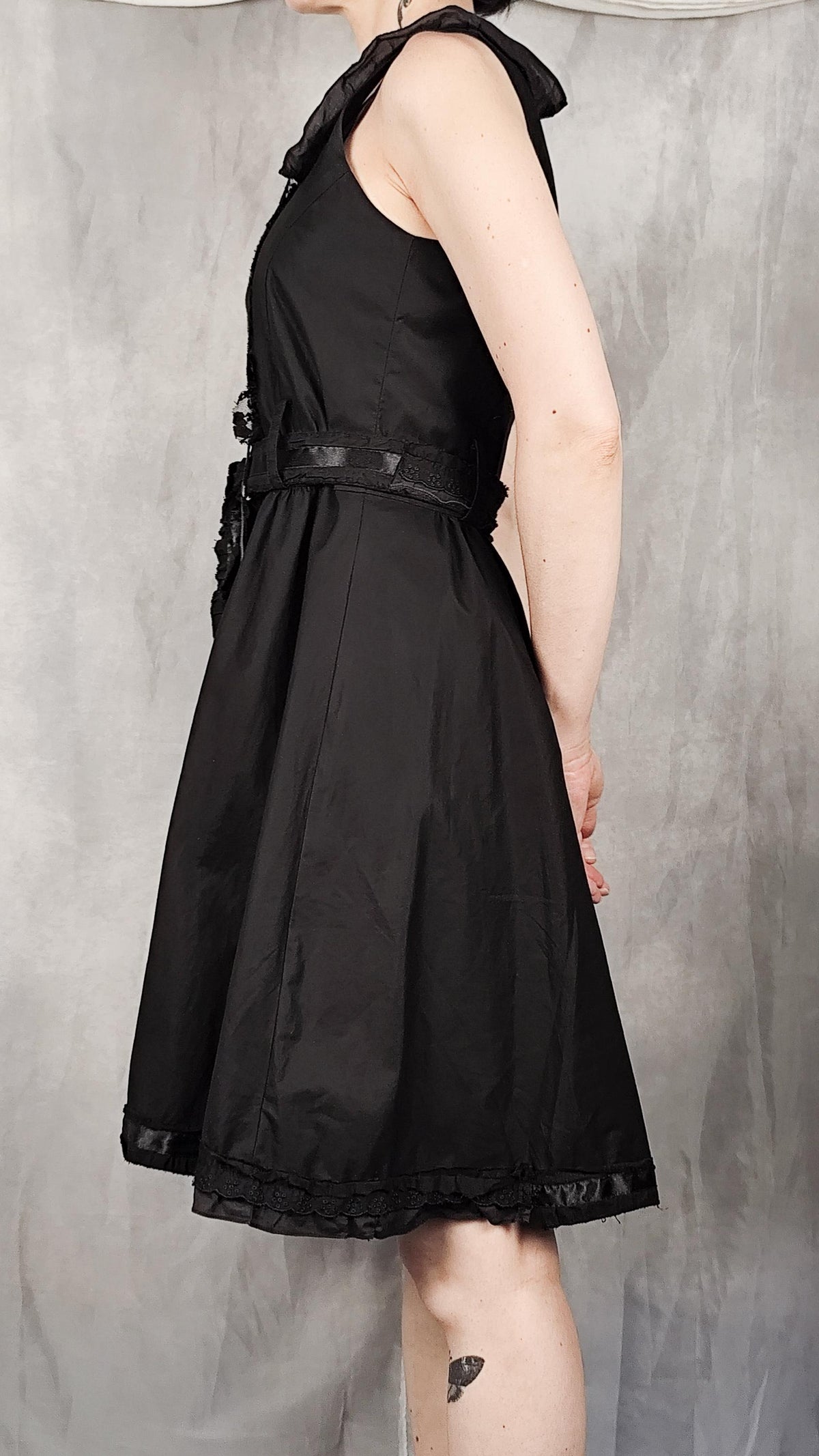 A sleeveless black dress with a ruffled collar, a matching black belt at the waist, and a flared skirt, shown on a model against a textured gray backdrop.