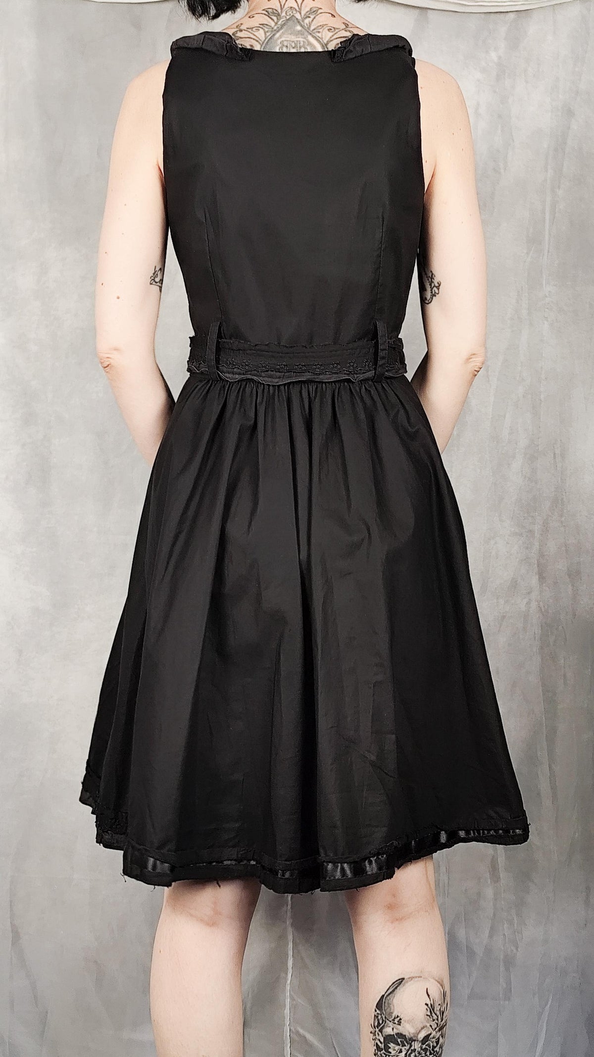 A sleeveless black A-line dress with a defined waist and a flared skirt, shown from the back on a model against a textured gray backdrop.