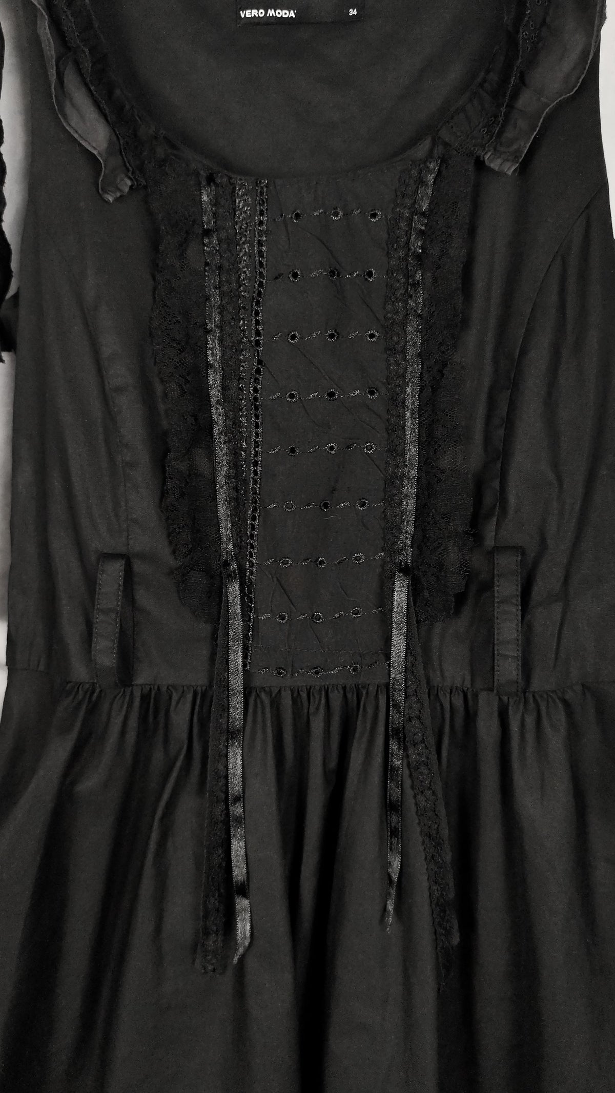 A close-up of a black dress featuring a decorative lace-up bodice with english embroidery details, sheer ruffled trim, and a gathered skirt.