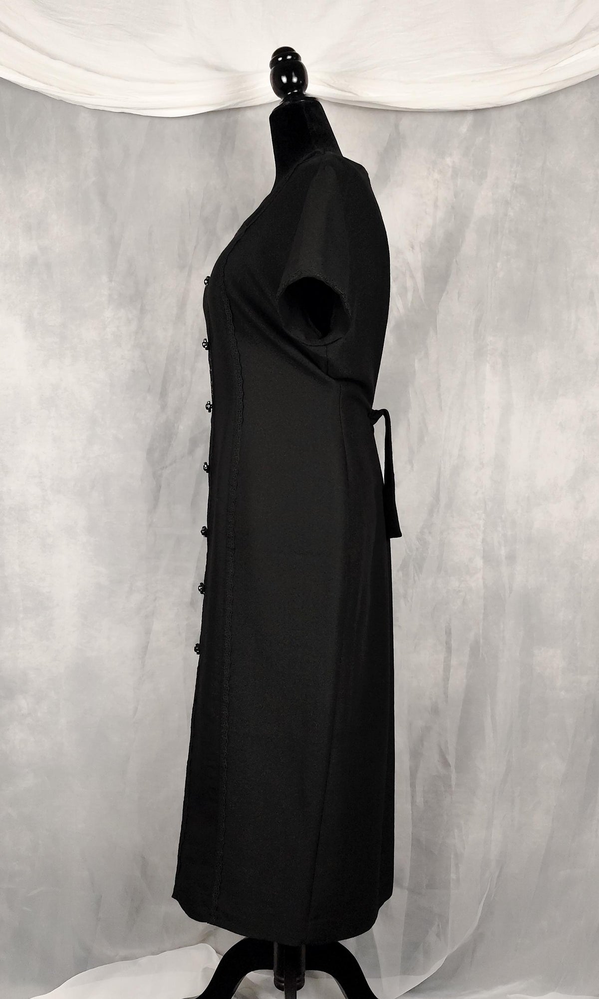 Side view of a black short-sleeve vamp midi dress displayed on a black mannequin against a gray draped fabric backdrop.