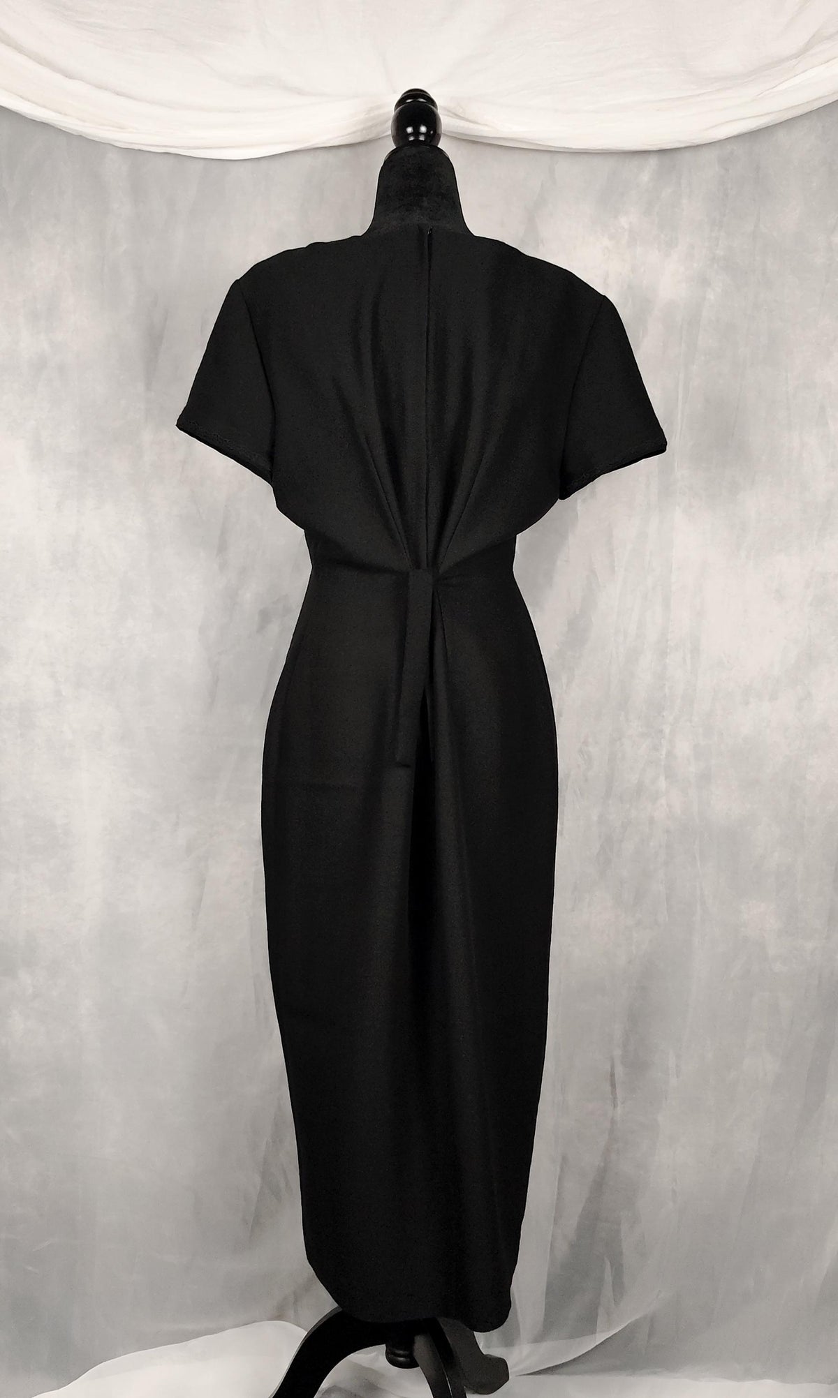 Back view of a black short-sleeve vamp midi dress, tied with a knot, displayed on a black mannequin against a gray draped fabric backdrop.