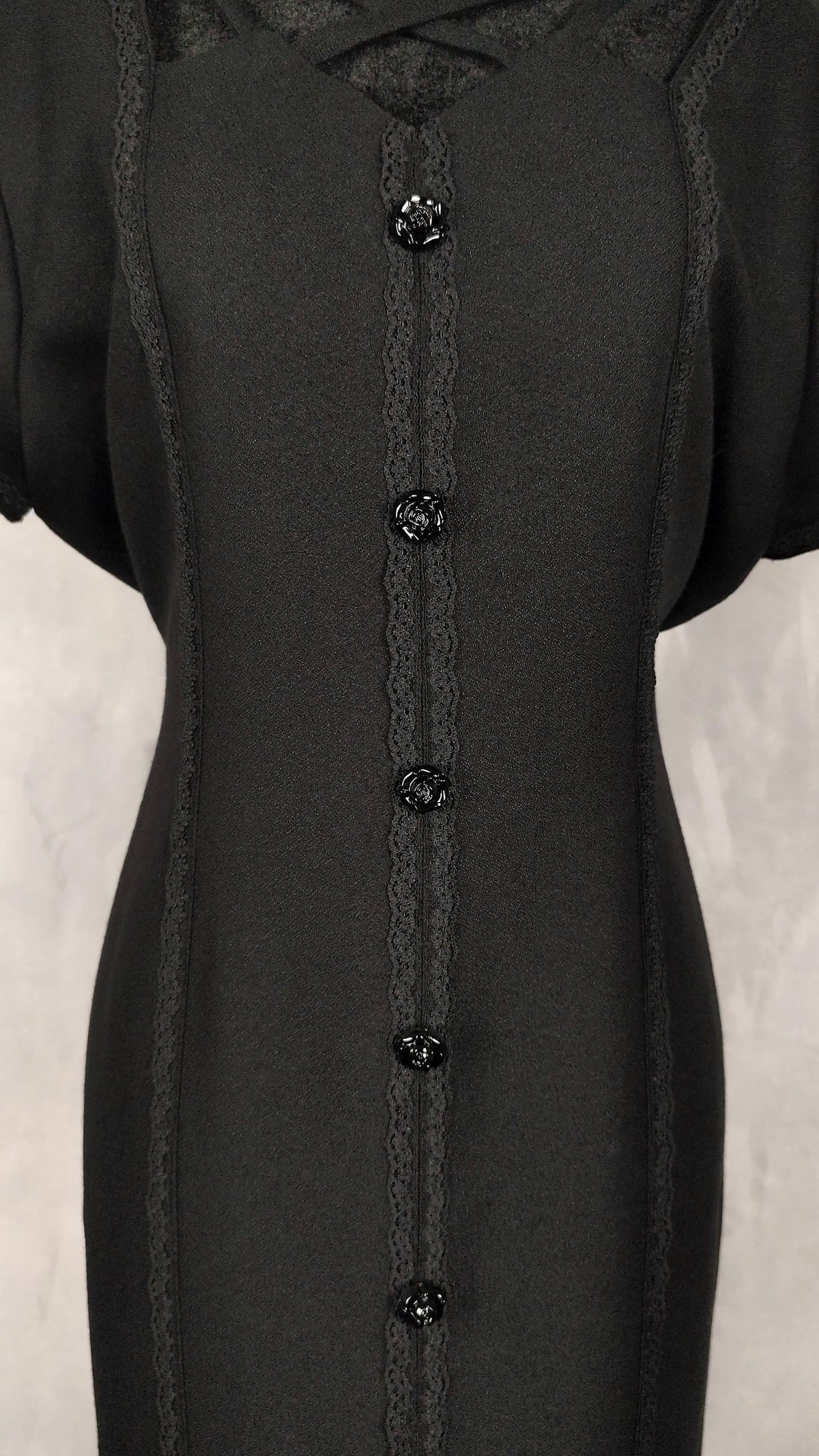 A close-up of a black short-sleeve midi dress featuring decorative lace trim, and a vertical row of black buttons down the center front.