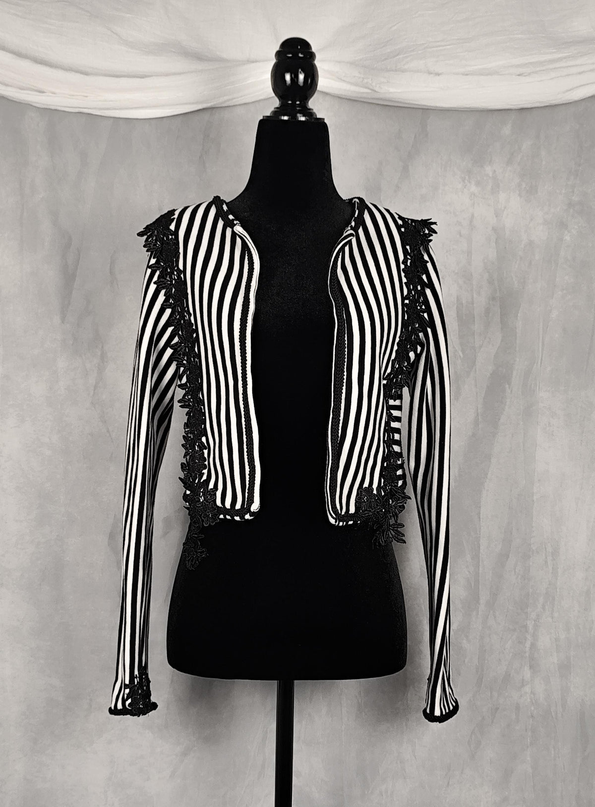 Front view of a black and white vertically striped cropped jacket with black lace trim, displayed on a black dress form mannequin against a textured gray backdrop.