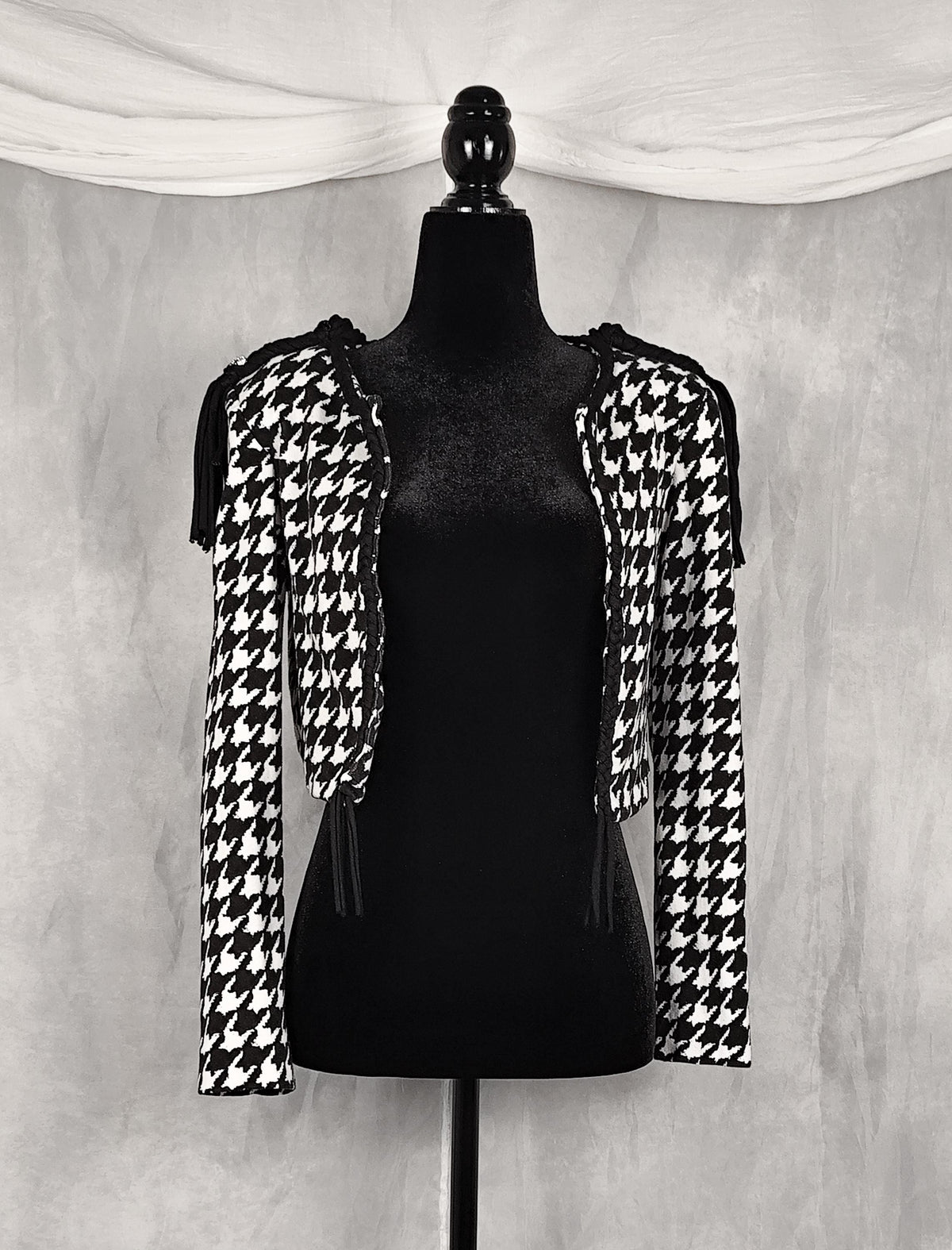 Front view of a black and white houndstooth cropped shrug jacket with shoulder cutouts and tassel ties, displayed on a black mannequin against a textured gray backdrop.