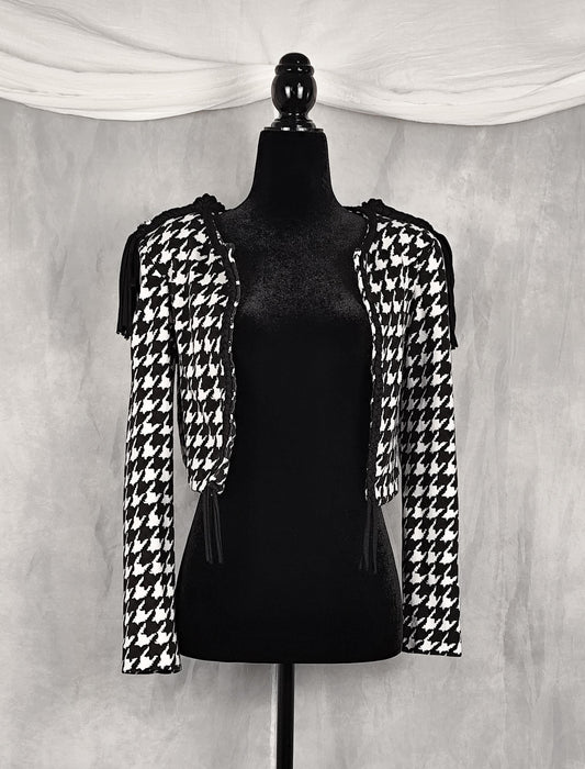 Front view of a black and white houndstooth cropped shrug jacket with shoulder cutouts and tassel ties, displayed on a black mannequin against a textured gray backdrop.