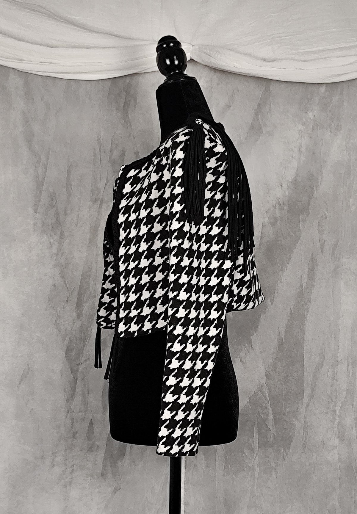 Side view of a black and white houndstooth cropped shrug jacket with shoulder cutouts and tassel ties, displayed on a black mannequin against a textured gray backdrop.