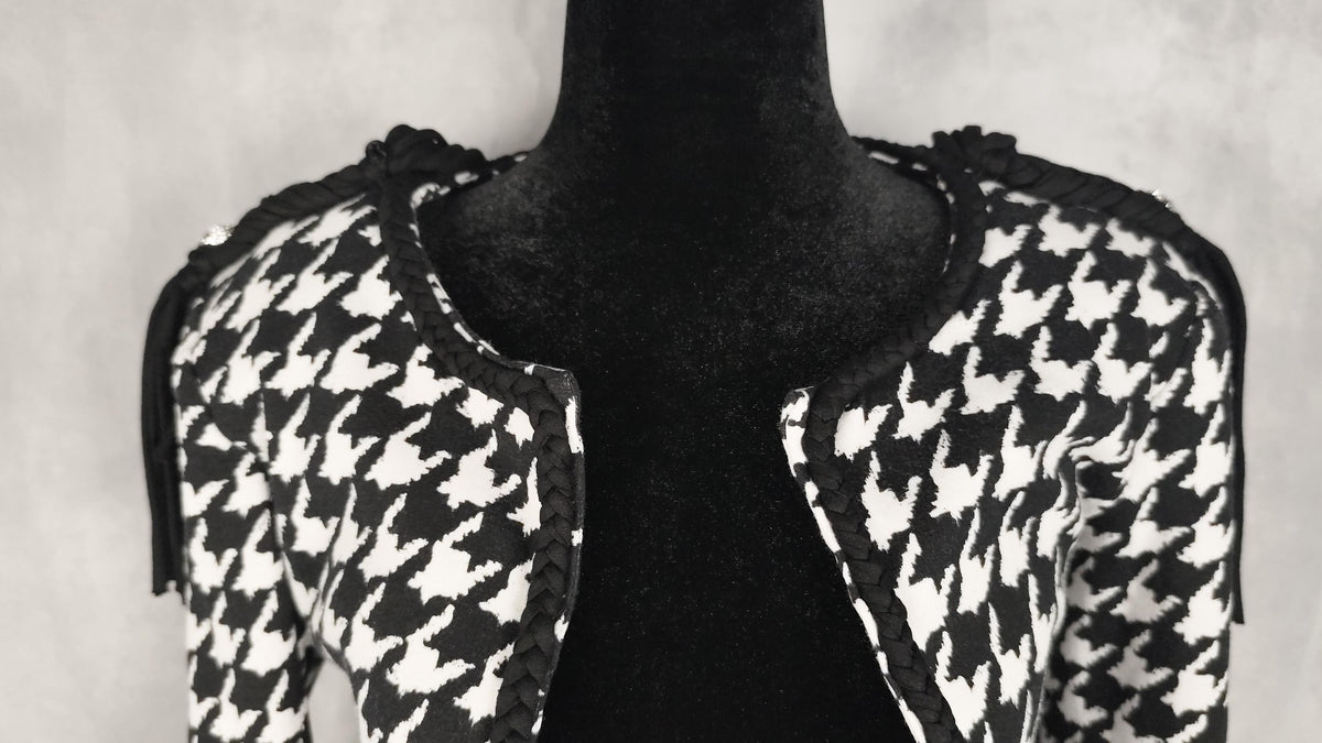 A black and white houndstooth jacket with black braid trim, displayed on a black mannequin torso against a textured gray background.