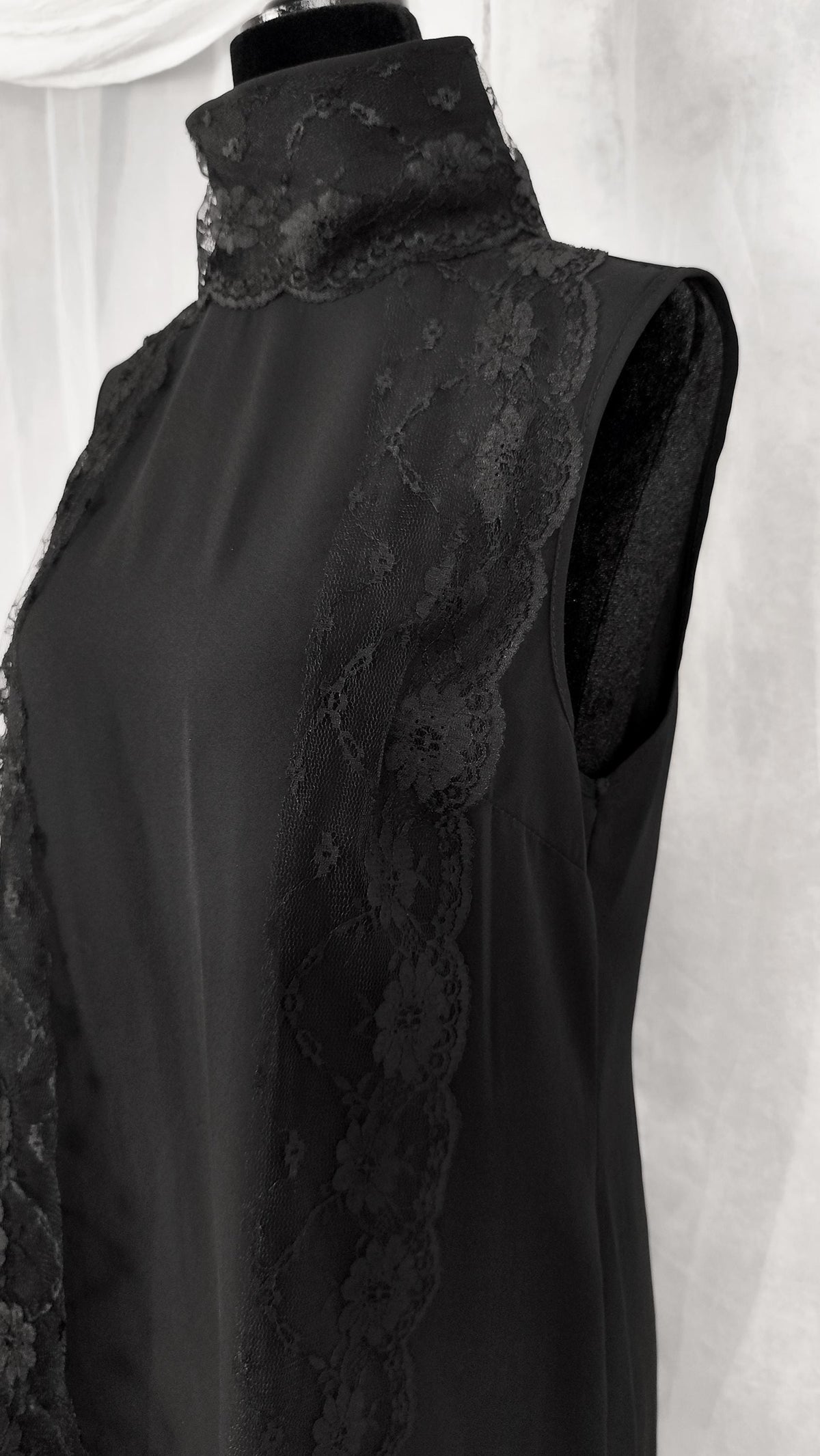 A black sleeveless high-neck tunic with sheer floral lace trim along the neckline and side panels, displayed on a black mannequin torso against a light draped fabric background.