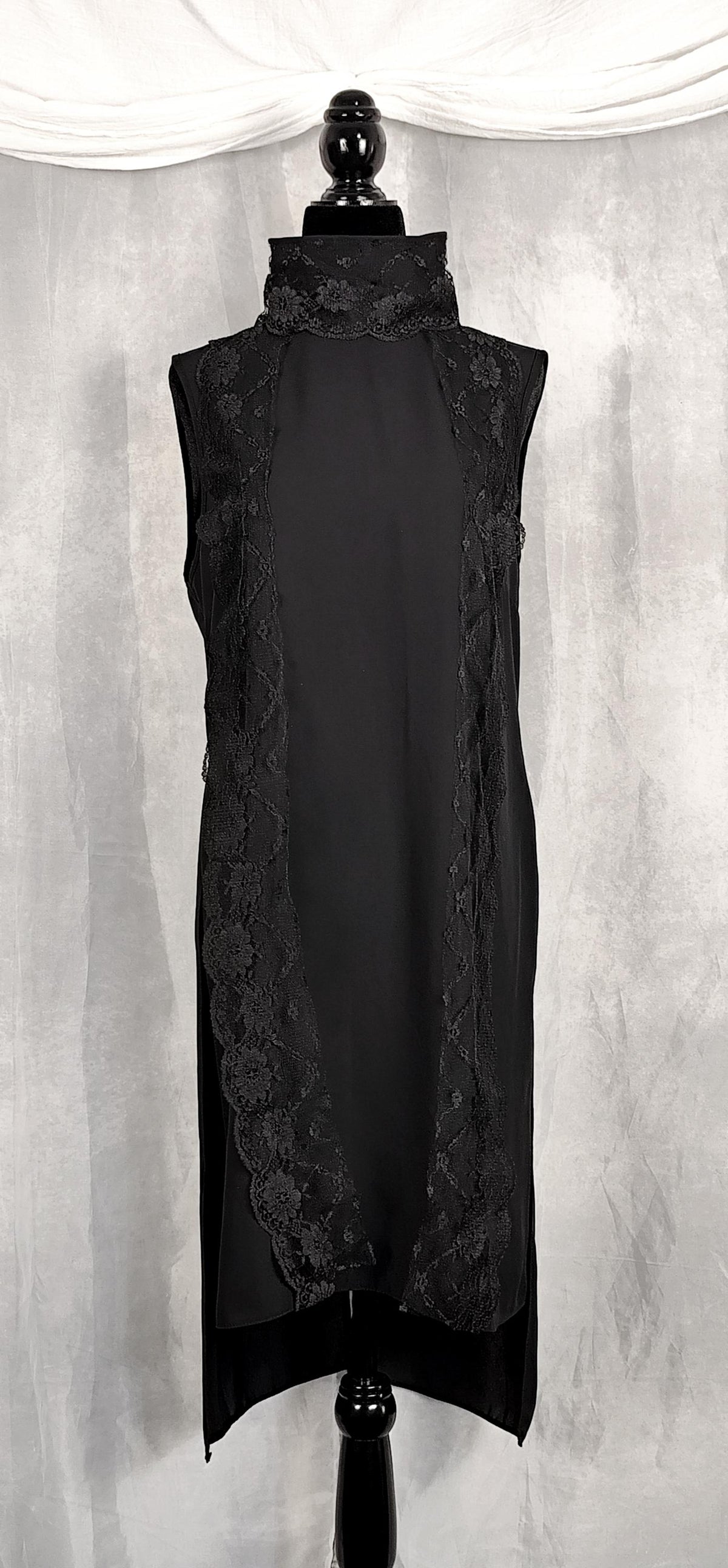 Front view of a sleeveless black long tunic with a high mock neck trimmed with lace and sheer lace side panels, displayed on a black mannequin against a gray draped fabric backdrop.