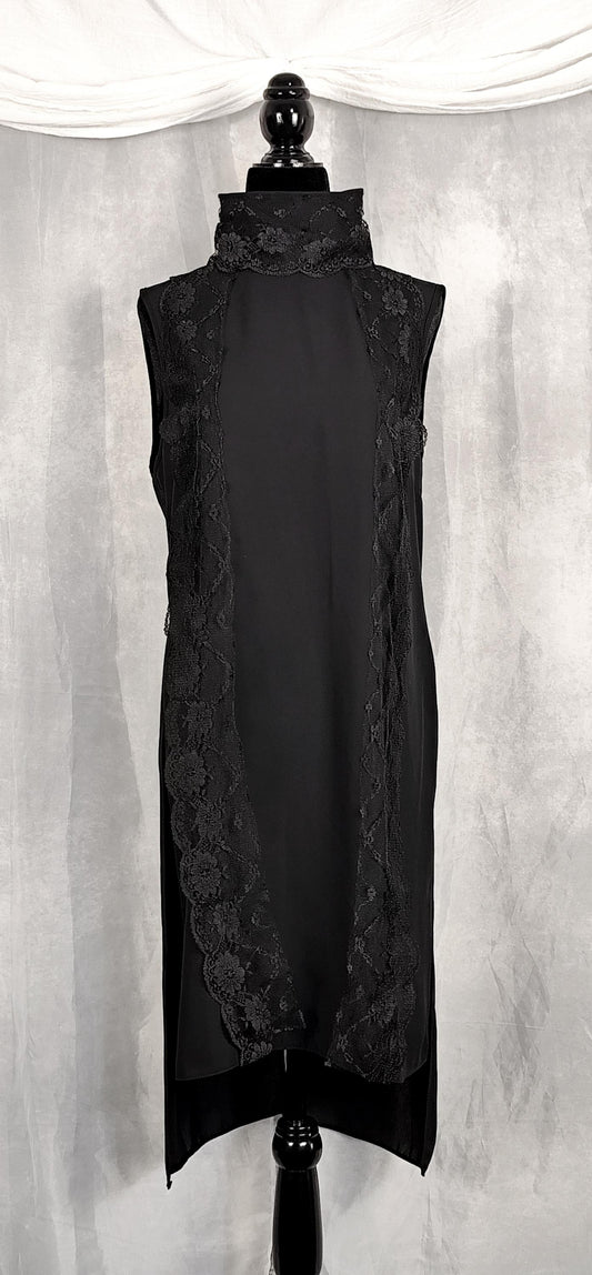 Front view of a sleeveless black long tunic with a high mock neck trimmed with lace and sheer lace side panels, displayed on a black mannequin against a gray draped fabric backdrop.