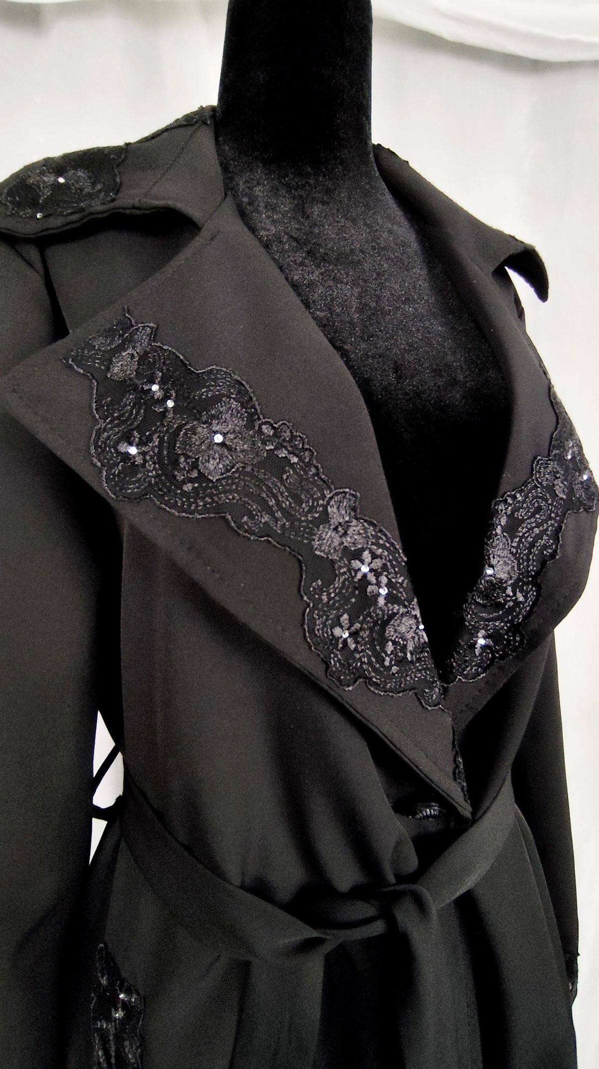 A black wrap-style trench coat displayed on a black mannequin torso, featuring wide lapels with black lace trim and sequined detailing, photographed against a light fabric backdrop.