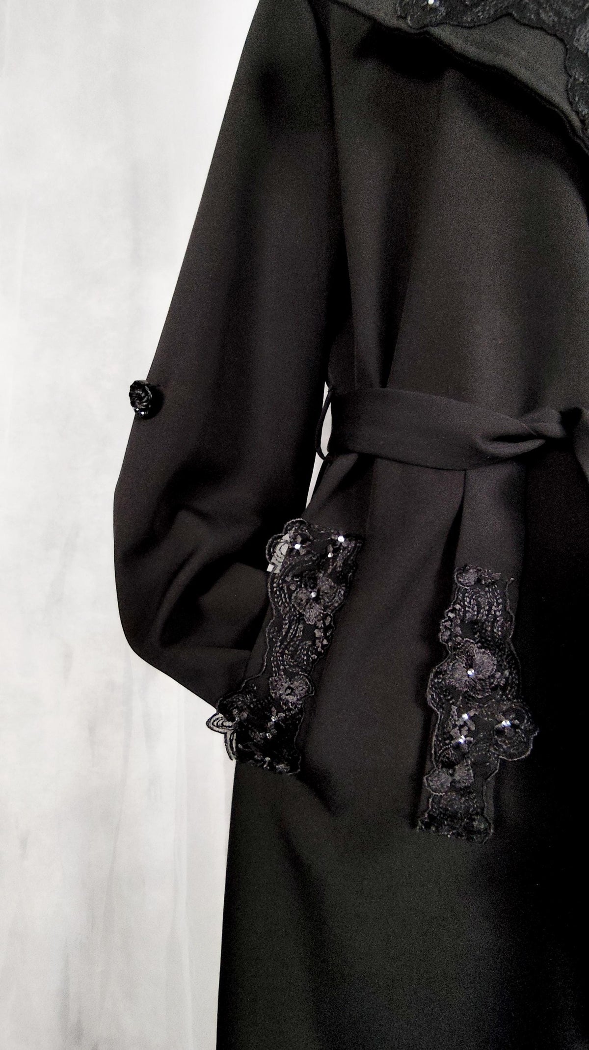 A close-up of a black belted trench coat featuring intricate black lace appliques on the cuffs and pocket flaps, set against a light gray textured background.