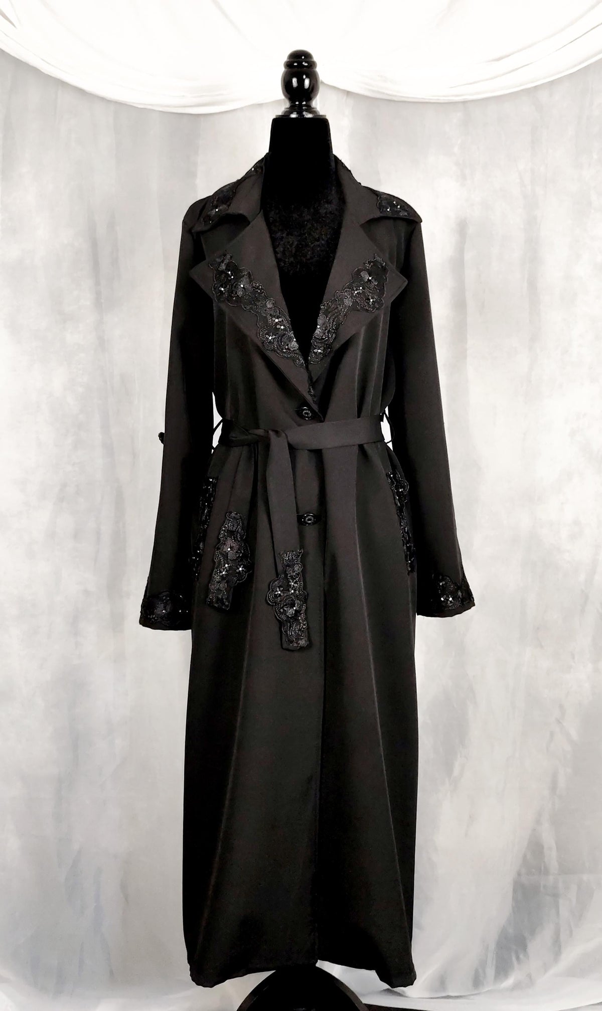 Front view of a long black trench coat with embroidered floral details on the lapels, cuffs, and waist belt, displayed on a black mannequin against a draped white fabric backdrop.
