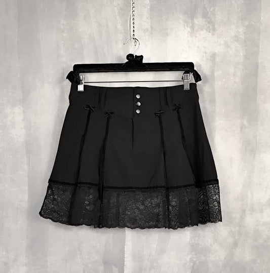 Front view of a black pleated mini skirt with a lace hem and three-button waistband, hanging on a black velvet hanger against a textured gray wall.