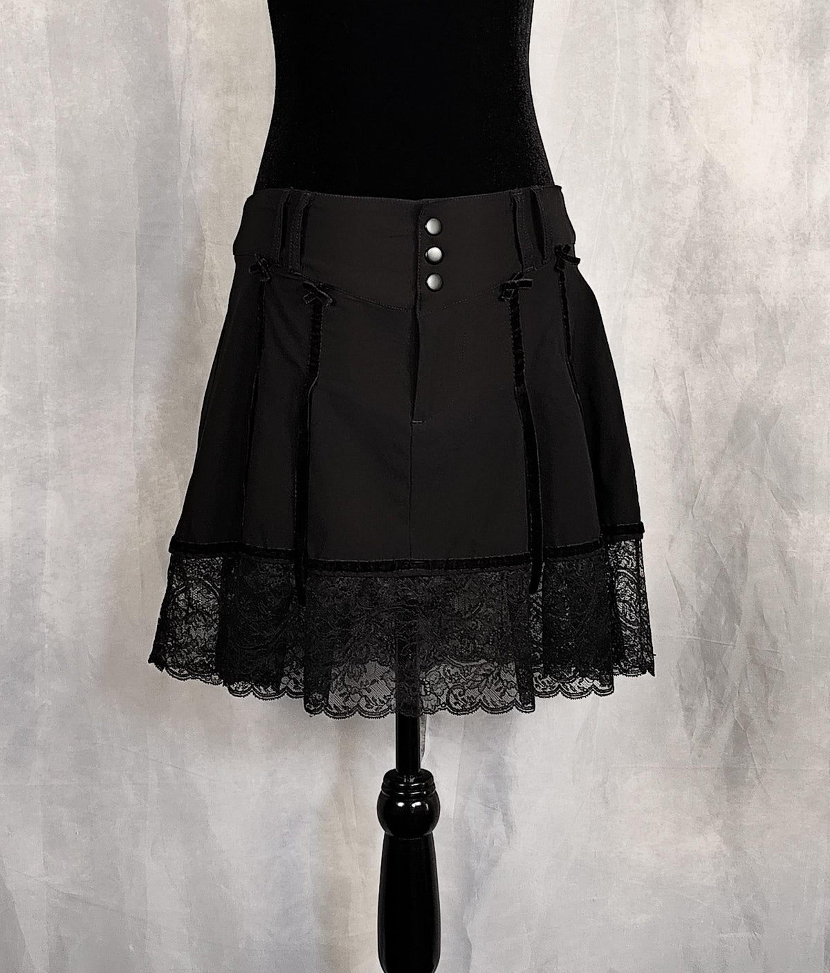 Front view of a black pleated skirt with a lace hem and three front buttons, displayed on a black mannequin torso against a textured gray backdrop.