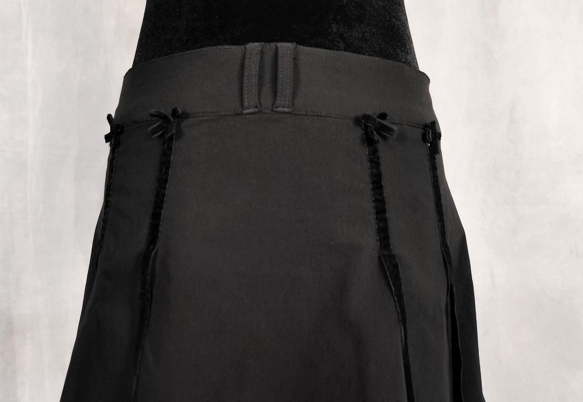 A close-up of a black pleated skirt with decorative velvet bows details at the waist and velvet ribbon, displayed on a mannequin against a light gray background.