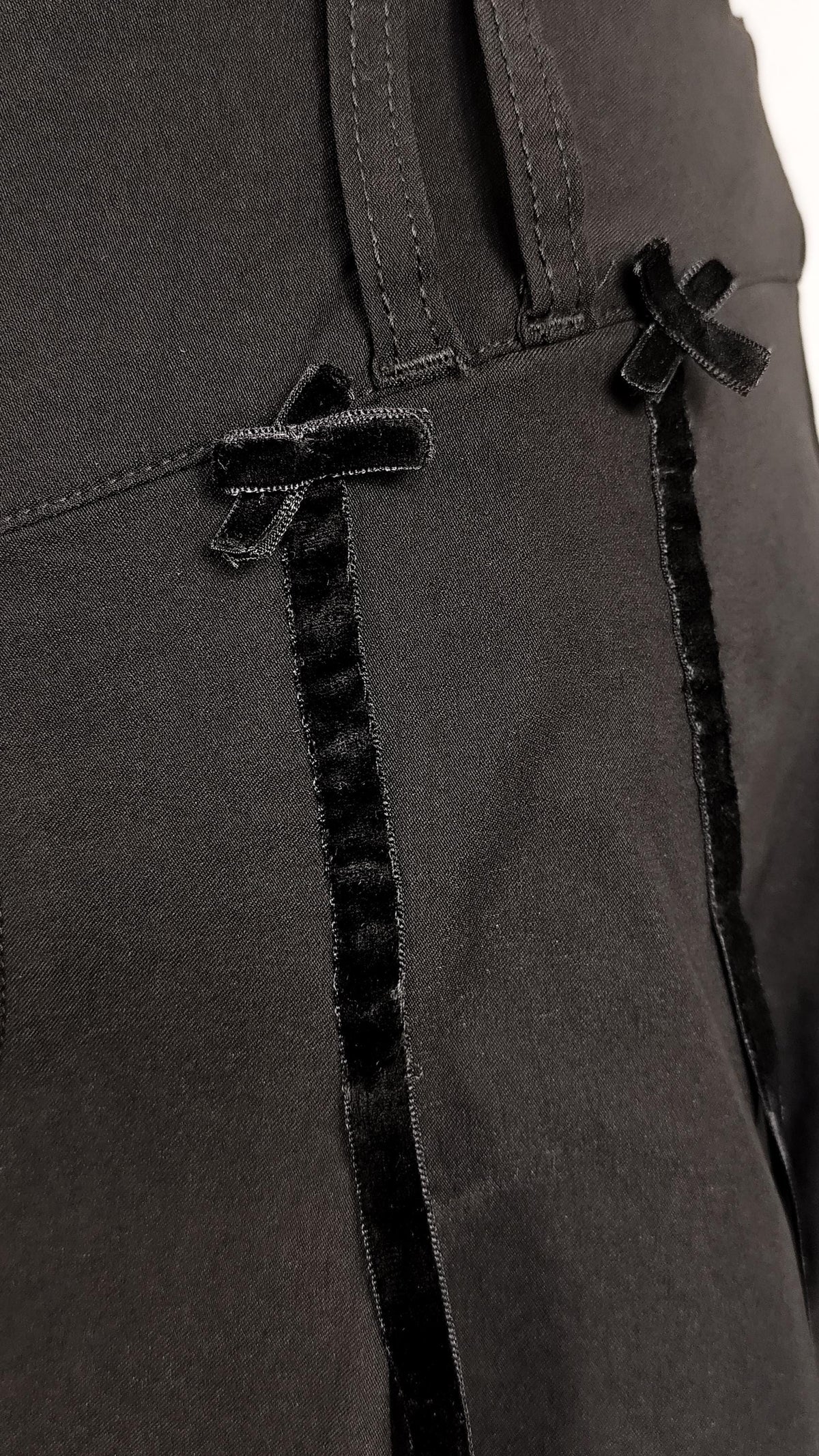 Close-up of a black skirt featuring vertical black velvet ribbon trims with bow details, set against a dark fabric background.
