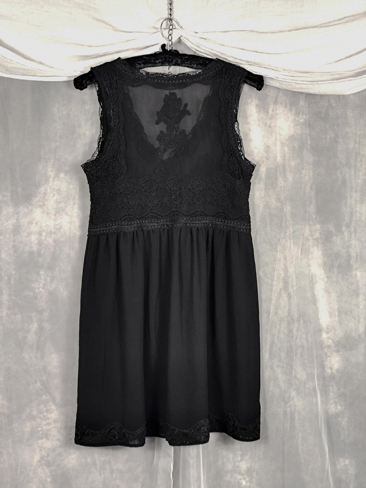 Back view of a black sleeveless nightgown with lace trim and a sheer floral back panel, hanging on a black hanger against a textured gray backdrop with draped white fabric at the top.