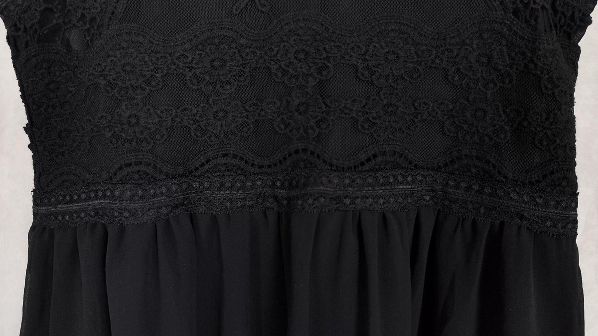 Close-up of a black midi dress featuring a sheer floral lace bodice overlay with a scalloped edge and a gathered black fabric skirt below.