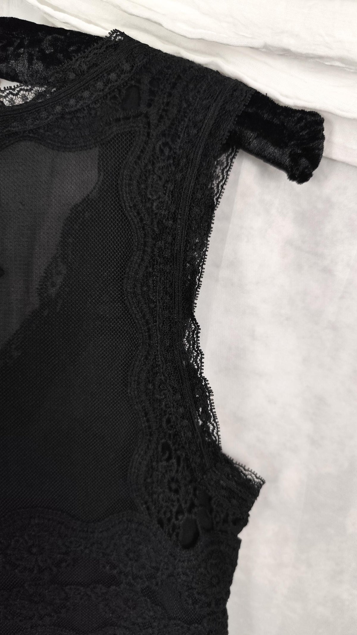 Close-up of a black lace and mesh sleeveless midi dress detail, showing scalloped lace trim and sheer fabric on a light textured background.