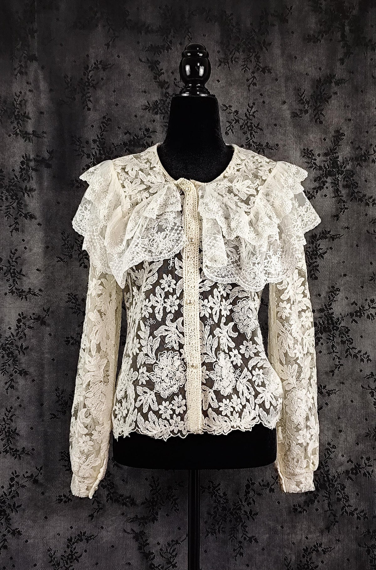 Front view of an intricate cream lace blouse with ruffled collar and a sheer floral lace bodice, displayed on a black mannequin torso against a dark textured backdrop.