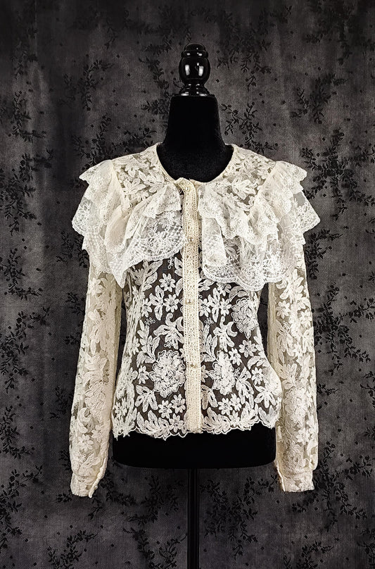 Front view of an intricate cream lace blouse with ruffled collar and a sheer floral lace bodice, displayed on a black mannequin torso against a dark textured backdrop.