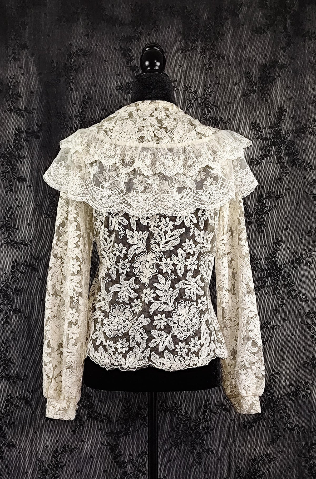Back view of an intricate cream lace blouse with a wide ruffled collar and long sleeves, displayed on a black mannequin torso against a dark patterned backdrop.