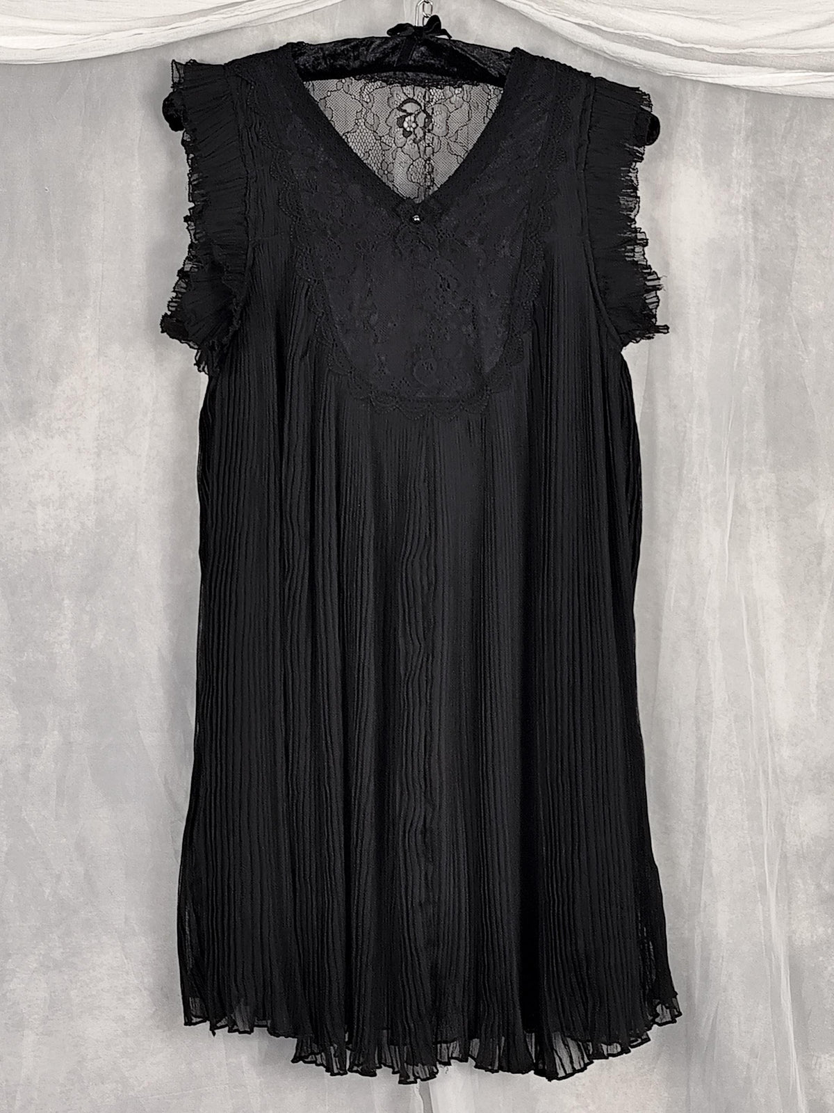 Front view black sleeveless pleated dress with a lace V-neck bodice and ruffled cap sleeves, hanging on a black hanger against a textured gray backdrop.