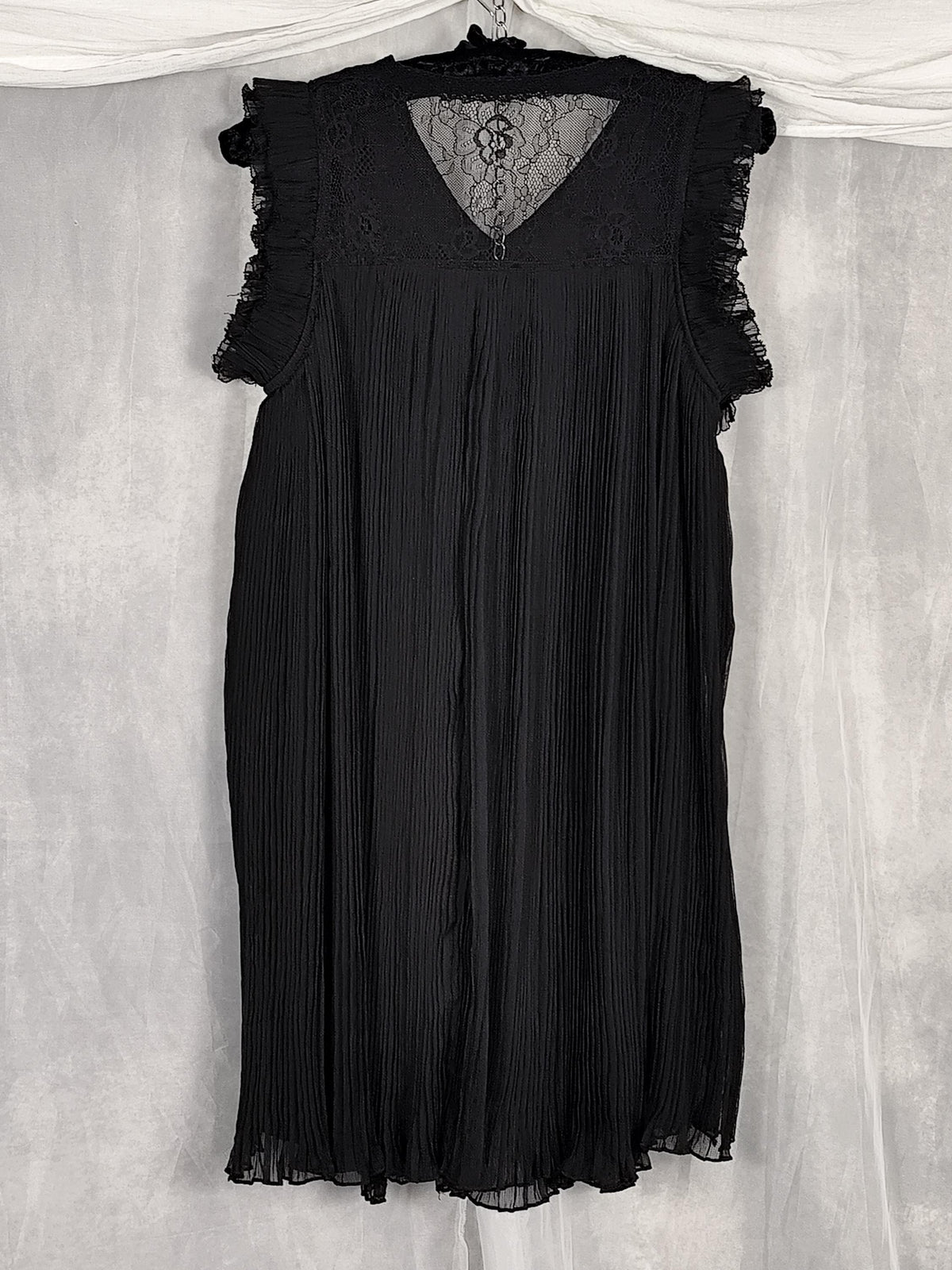 Back view of a black sleeveless pleated dress with ruffled cap sleeves and a sheer lace at the back and neck, hanging against a light gray fabric backdrop.