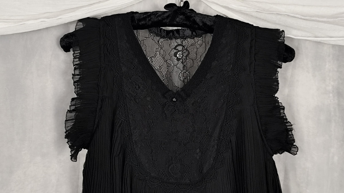 Close-up of a black sleeveless lace blouse with ruffled cap sleeves and a V-neckline, hanging on a black hanger against a draped white fabric backdrop.