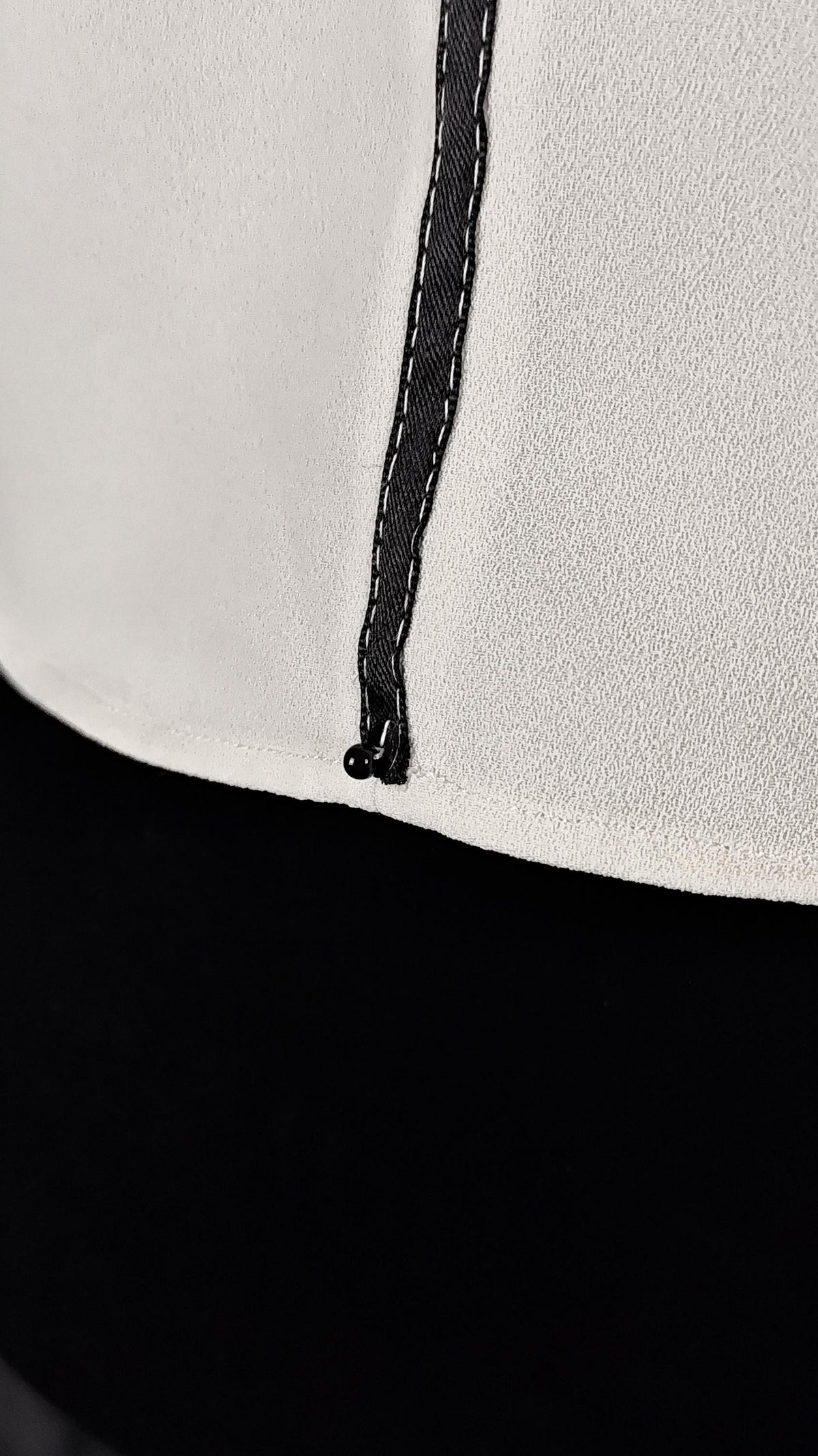 Close-up detail of a white textured garment with a black vertical stitched seam and a small black bead.