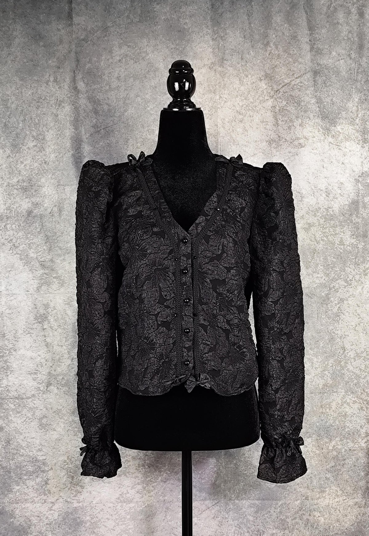 Front view of a black floral brocade blouse with black pearl-shaped buttons and braid trim.