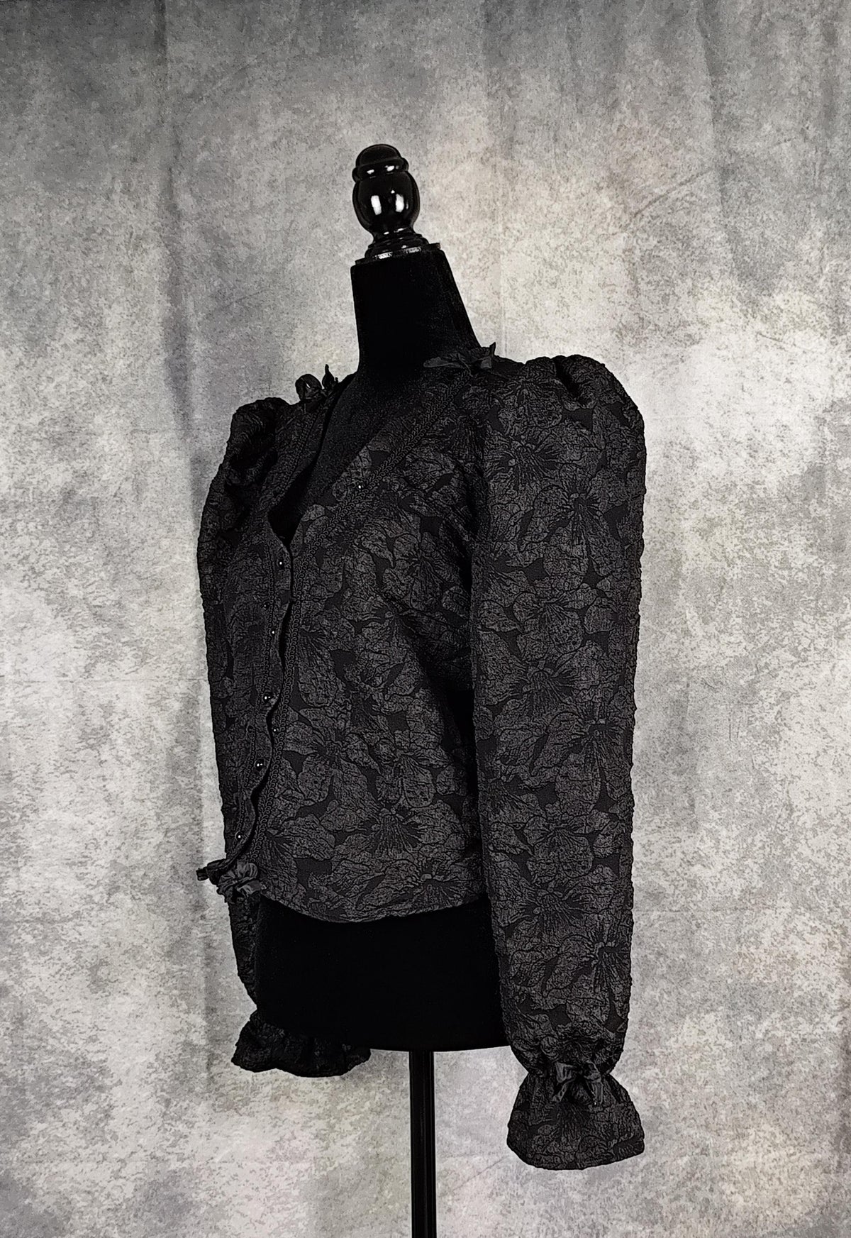 Side view of a black floral brocade blouse with black pearl-shaped buttons and braid trim.