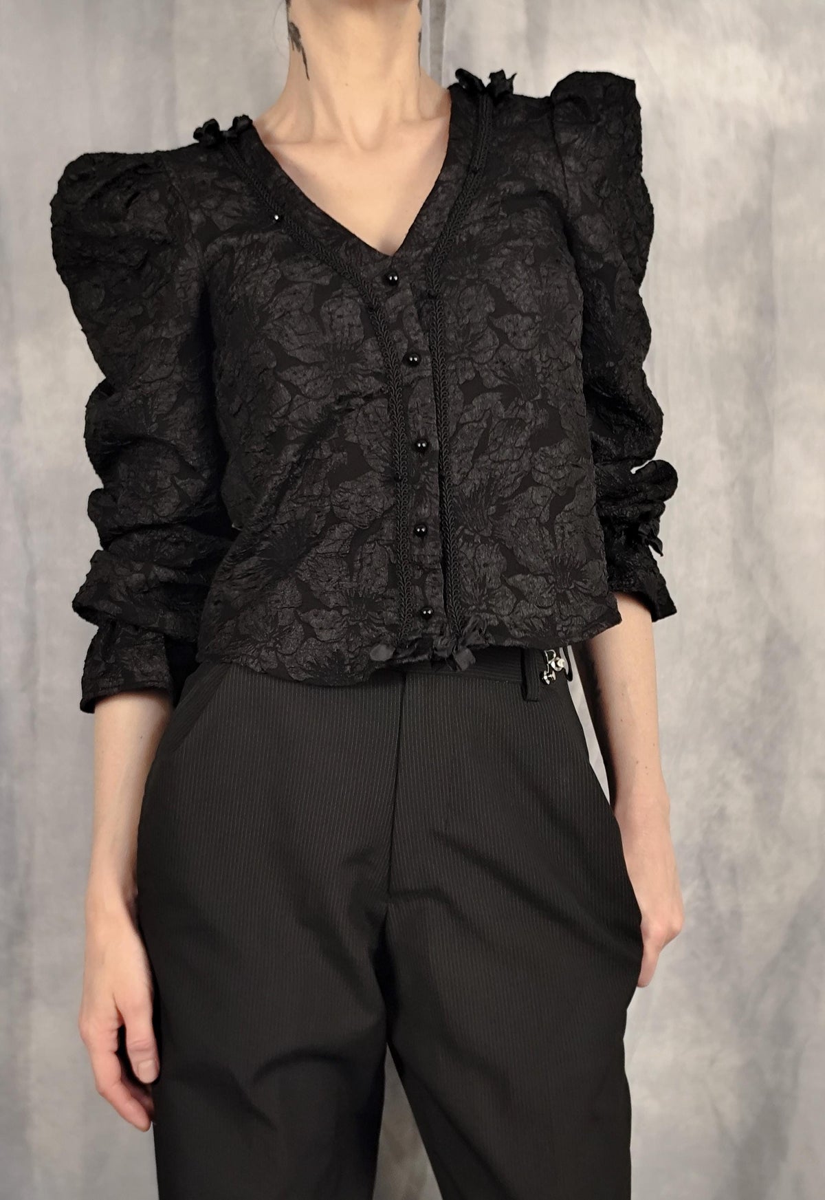 Model wearing a black gothic brocade blouse, with black pearl-shaped buttons, braid trim, bows and puff longsleeves.
