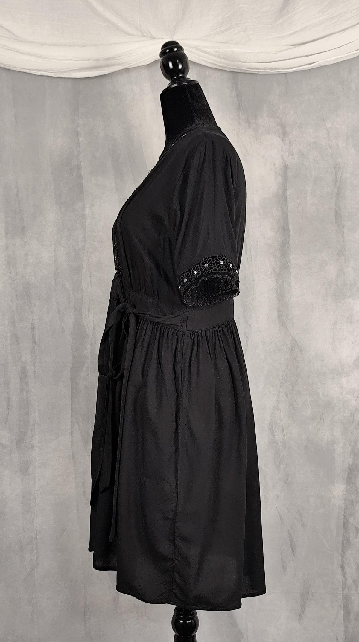 Side view of a knee high black short-sleeve wrap dress with decorative silver beaded trim along the V-neckline and sleeve cuffs, trimmed with velvet and tulle.
