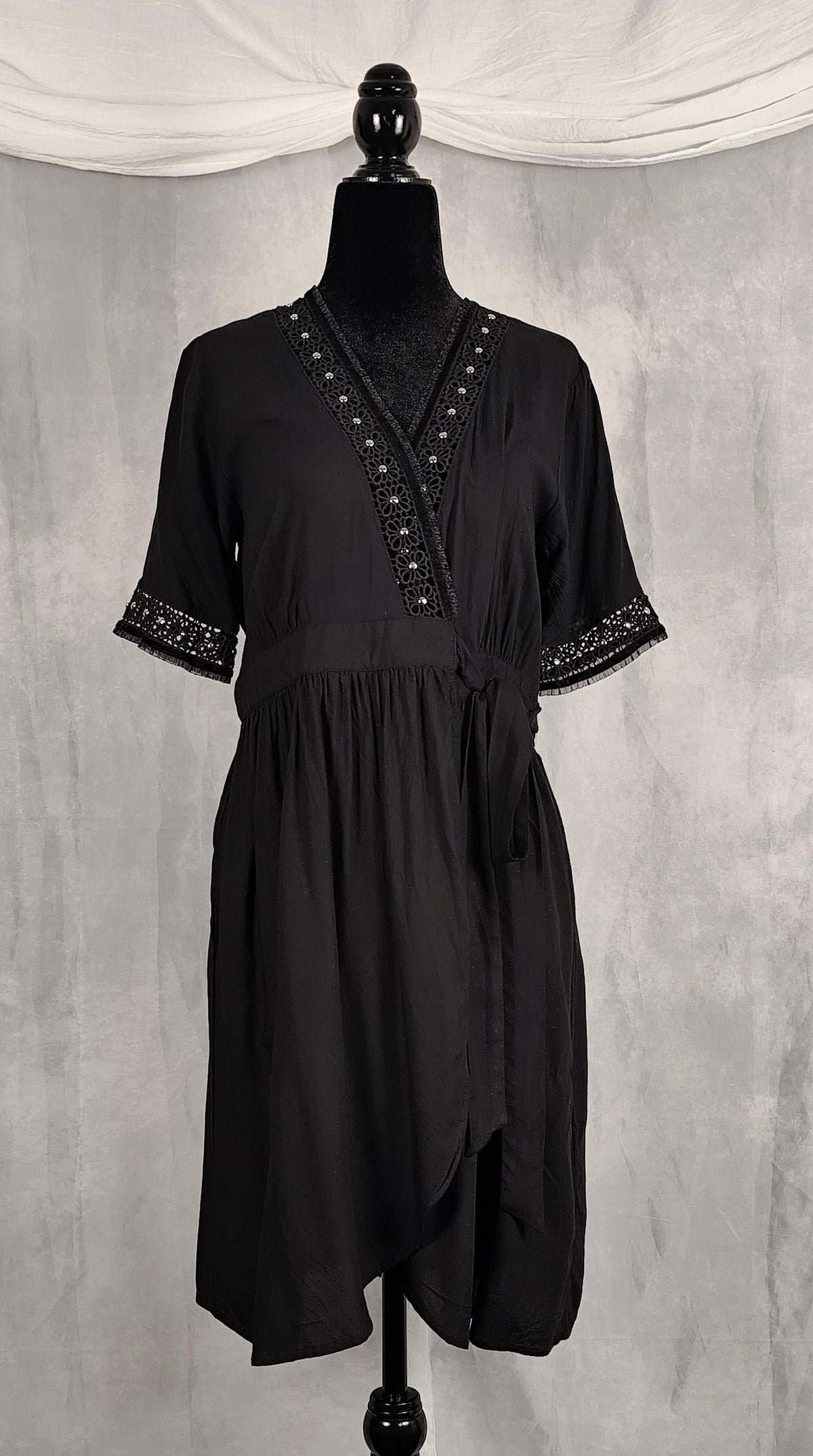 Front view of a knee high black short-sleeve wrap dress with decorative silver beaded trim along the V-neckline and sleeve cuffs, trimmed with velvet and tulle.