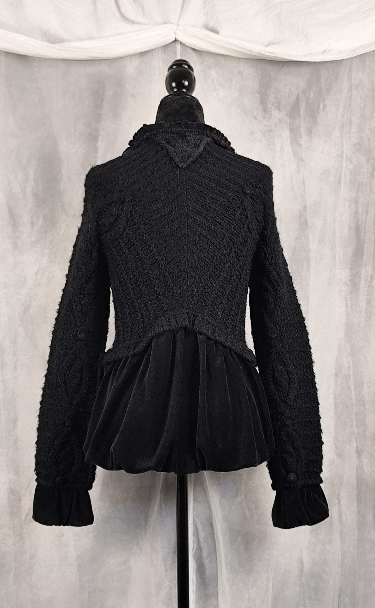 Back view of a black textured knit and velvet jacket displayed on a black mannequin torso against a gray draped fabric backdrop.
