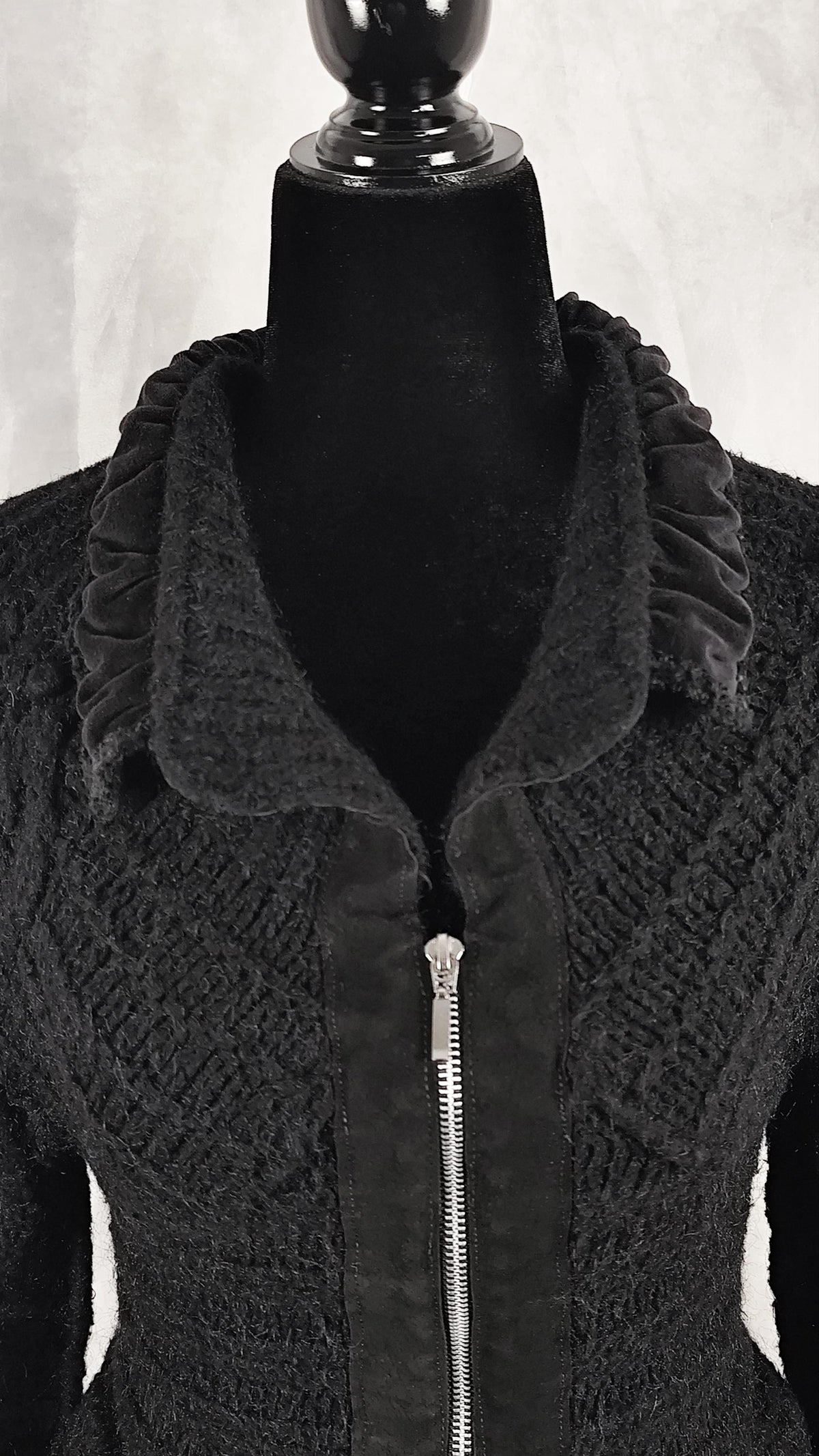 A black textured knit jacket with a ruffled velvet collar and a central silver zipper, displayed on a black mannequin torso against a light gray background.